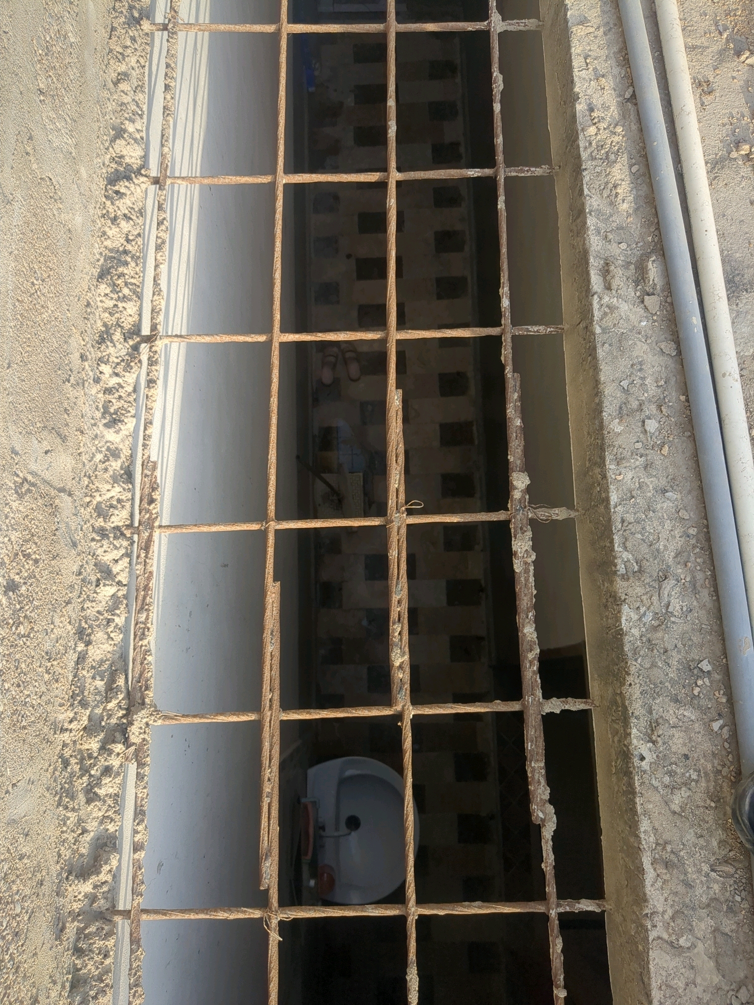 The image shows a close-up, high-angle view of a metal grate covering a drainage or utility channel. The grate is made of rusted rebar that appears to be twisted together. Below the grate, a white ceramic sink is visible, along with some patterned tiling on the floor of the channel. Two pairs of sandals are also present on the floor. To the right of the grate, two light-colored pipes are lying on the ground next to a concrete edge. The surrounding environment appears to be an outdoor urban setting, possibly a street or alleyway in Karachi, Pakistan. The overall scene suggests a neglected or disused area.