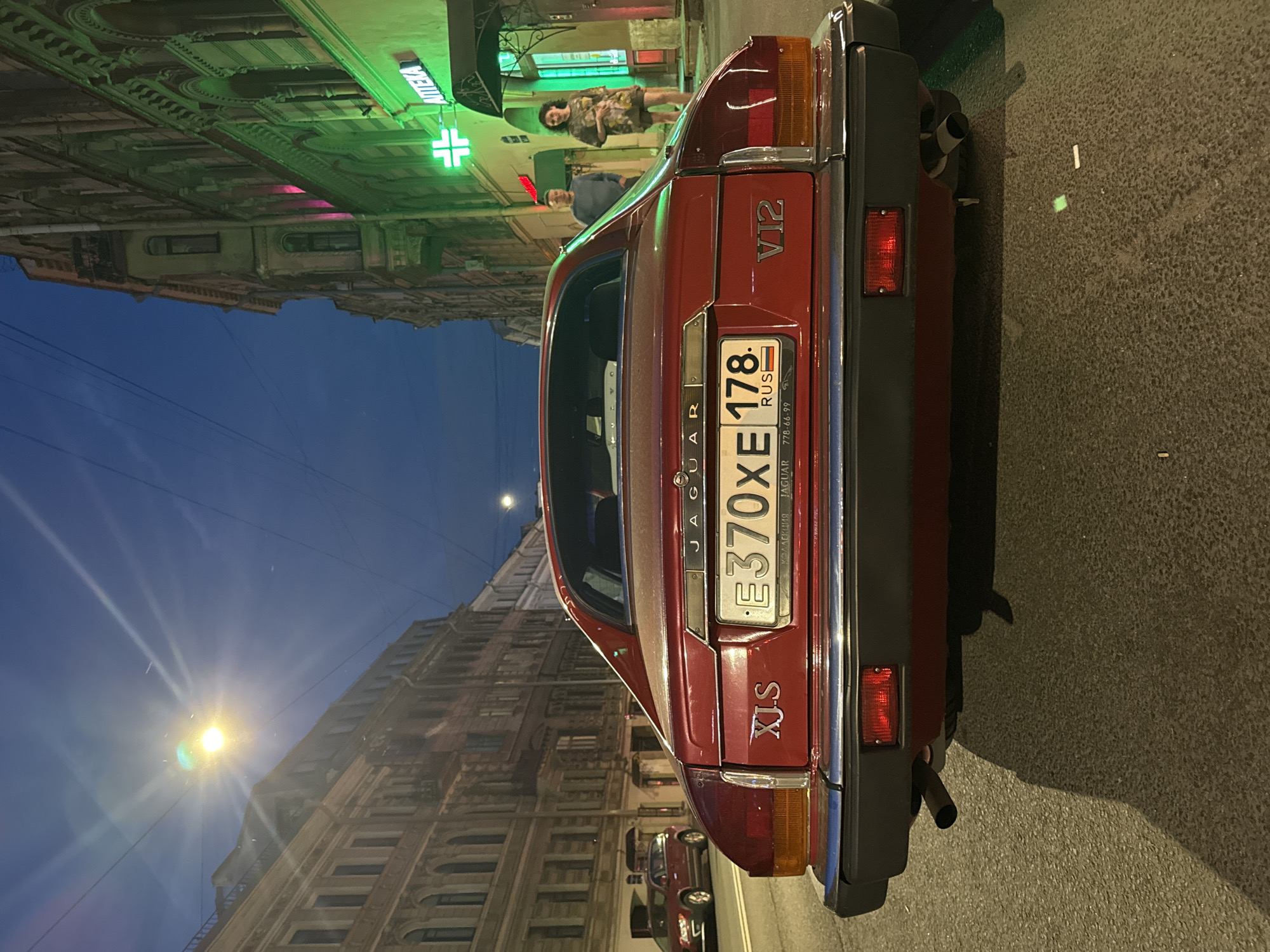 A red 1988 Jaguar XJ-S V12 is parked on a cobblestone street at dusk in Saint Petersburg, Russia. The car's rear is facing the camera, and its license plate reads "E370XE 178 RUS". The street is lined with historic European-style buildings, one of which has a lit green pharmacy sign with a cross. Two people, a man and a woman, are walking away from the camera on the sidewalk in front of the pharmacy. The sky is a deep blue, and a streetlamp illuminates the scene, casting long shadows on the ground.