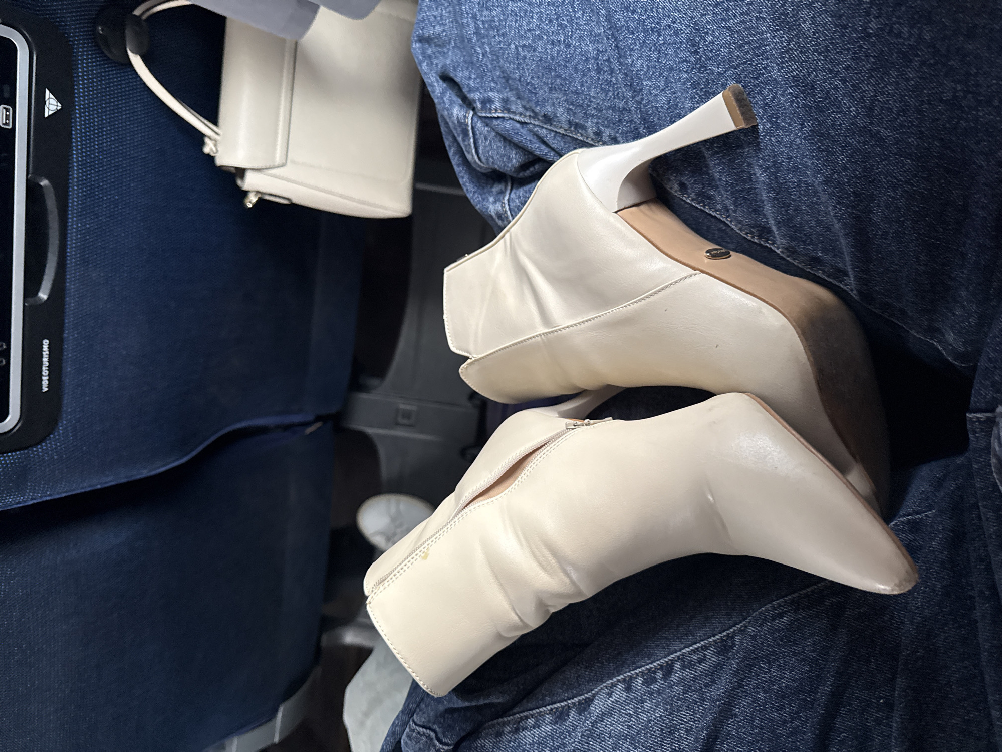 The image shows a pair of off-white heeled ankle boots resting on a person's lap. The person is wearing denim jeans. To the left, a blue airplane seat is visible. A small white handbag is partially visible. The location is not apparent from the image and cannot be inferred.