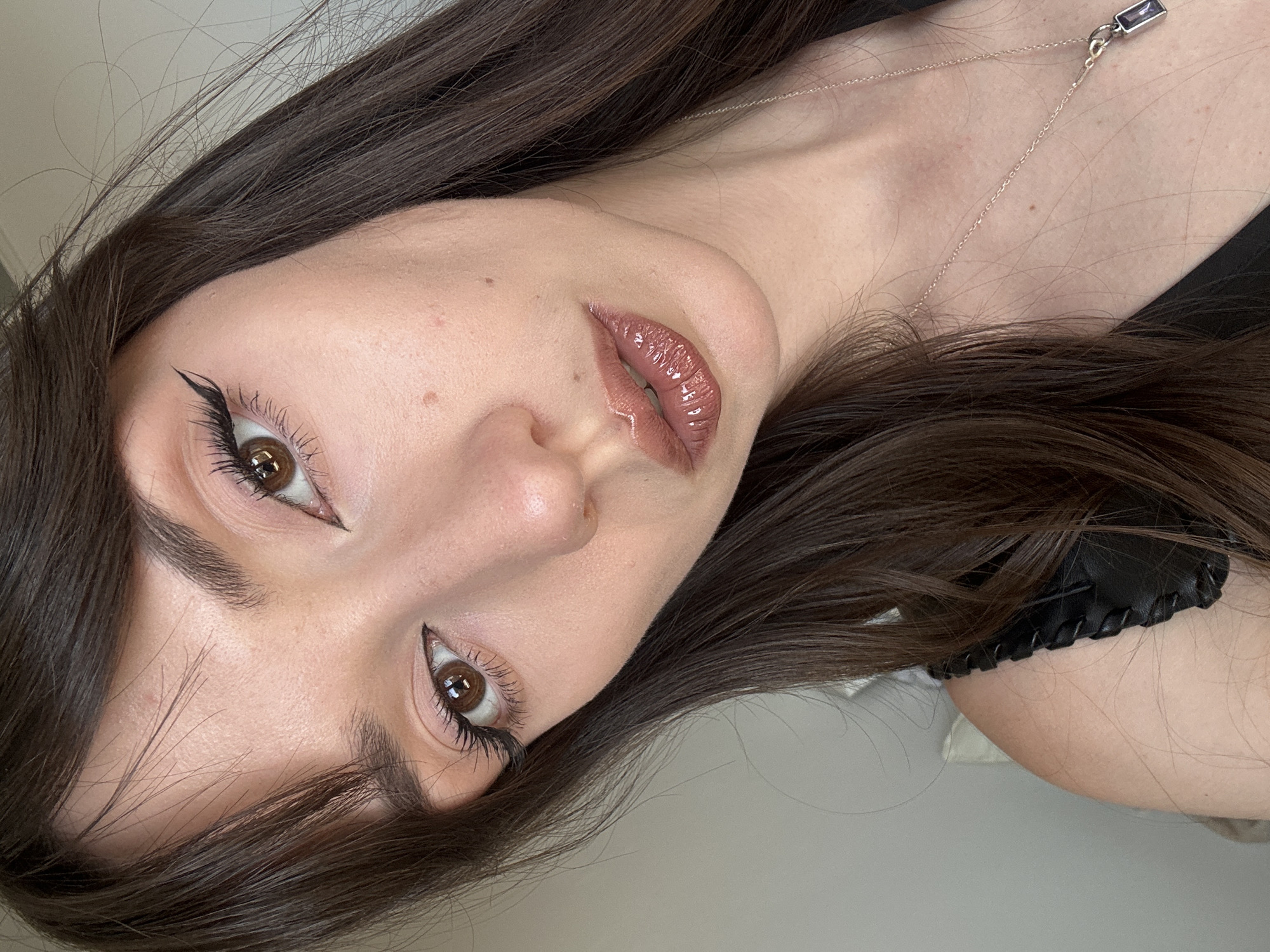 This is a close-up, overhead, tilted portrait of a young woman with long, dark brown hair. She is wearing a black top with a decorative stitched detail on the strap and a delicate silver necklace with a small, rectangular, dark purple pendant. Her makeup includes prominent winged eyeliner and voluminous mascara, with a glossy, neutral-toned lipstick. The lighting suggests it might be daytime, possibly indoors given the neutral background. Her expression is neutral or slightly pensive. There are no visible text elements in the image. The context is Belgrade, Serbia.