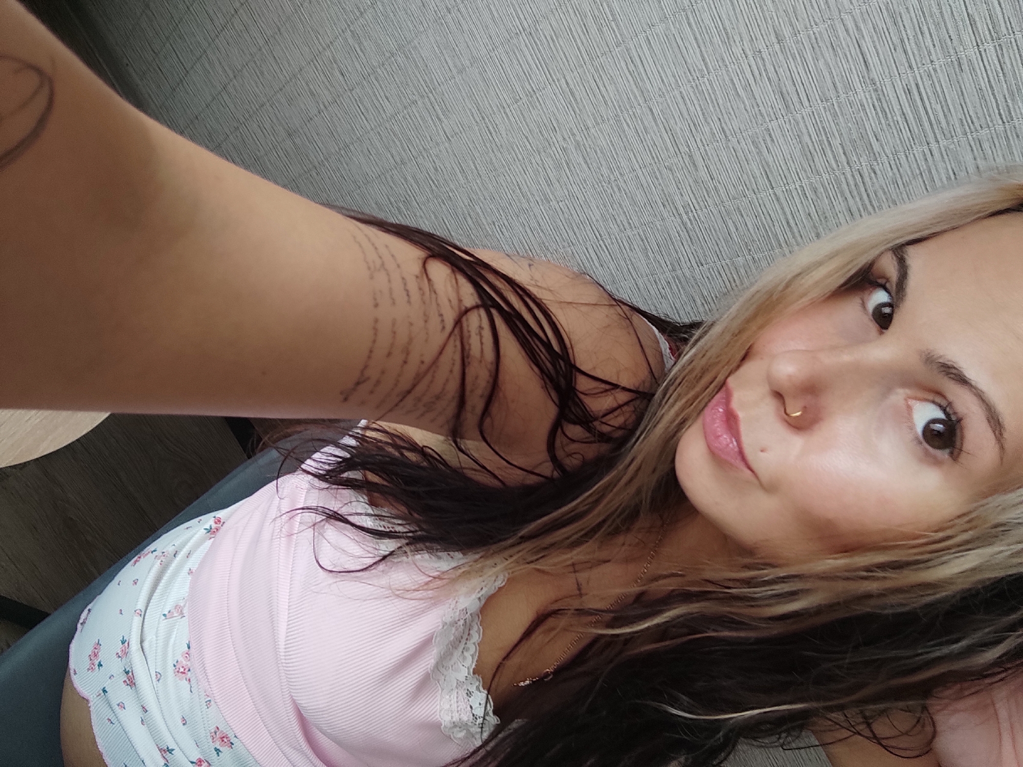 A woman is taking a selfie. She has blonde and dark brown hair, a nose ring, and light-colored skin. She is wearing a pink top with a floral-patterned bottom. The woman's arm is in the frame, showing a tattoo. The background is a textured wall. The location and city of Chaikovsky, Russia cannot be determined from the image.