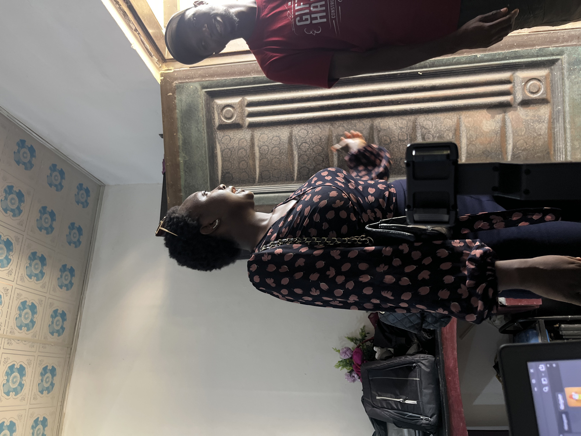 The image depicts two individuals in what appears to be an indoor setting in Gura Top, Nigeria. In the foreground, a woman with dark, curly hair and sunglasses perched on her head is looking upwards and to her left, with her mouth slightly open as if speaking or laughing. She is wearing a dark, long-sleeved blouse with a pattern of small, light-colored, oval shapes. A black purse with a gold chain strap is draped over her shoulder. Behind her, and slightly out of focus, is a large, dark-colored door with decorative molding.
Above the woman and partially obscured is a man wearing a red t-shirt and a black cap. He is looking down and smiling. The t-shirt has visible white text that appears to read "GIFTED" and "HANDS" followed by other, less legible words, possibly indicating "CONSTRUCTION."
The setting includes a wall with a tiled pattern featuring blue circular designs. To the right of the woman, there are bags and a glimpse of a tablet screen displaying an interface with icons. The lighting suggests it is daytime. The overall mood seems casual and possibly social or a behind-the-scenes moment.