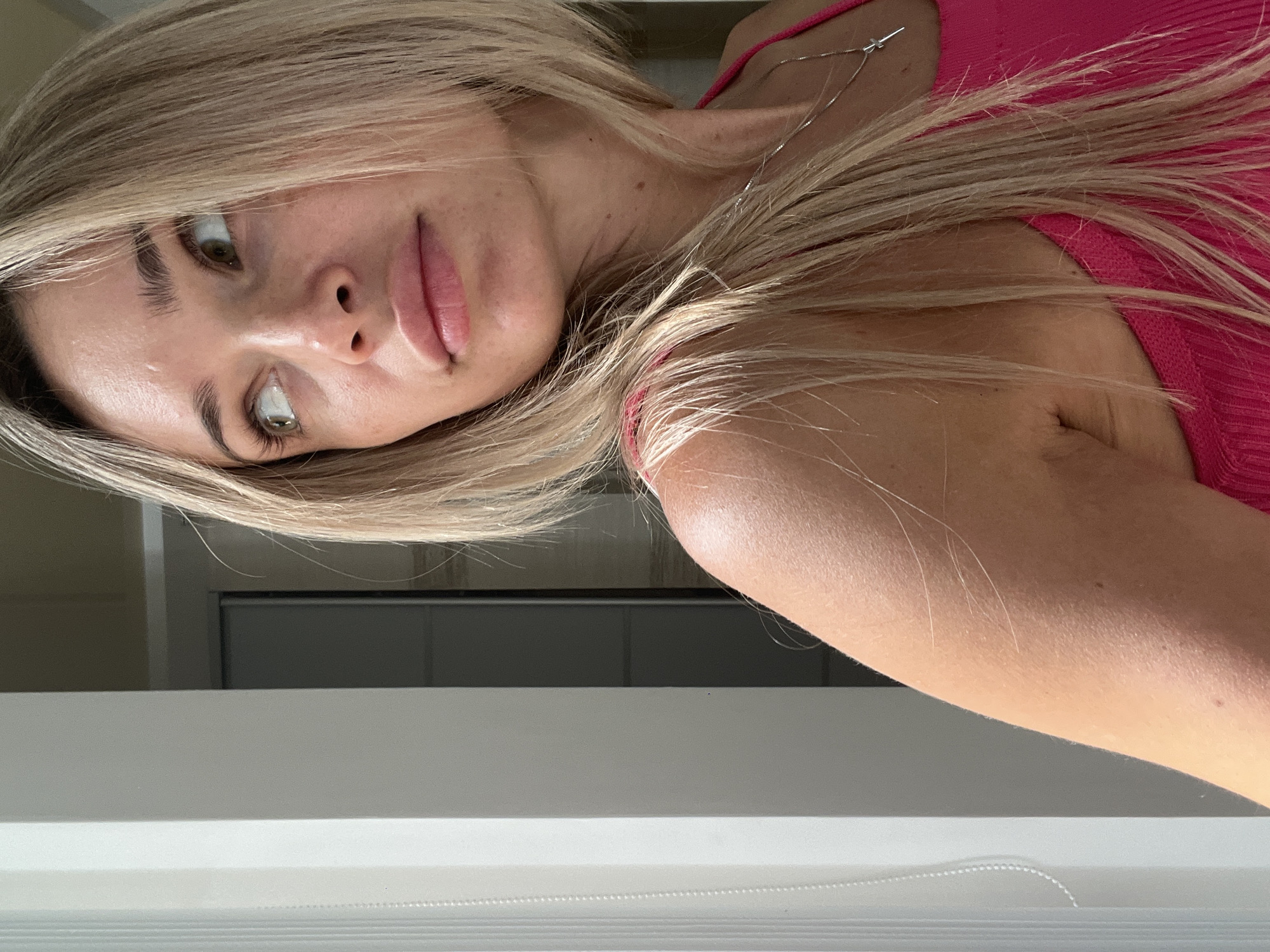 A woman with blonde hair and green eyes is shown from the chest up, looking to her left. She is wearing a pink tank top and a silver necklace with a cross pendant. The background appears to be indoors, with a white vertical blind on the left and a doorway with a dark door visible on the right. The lighting suggests it is daytime, possibly near a window.