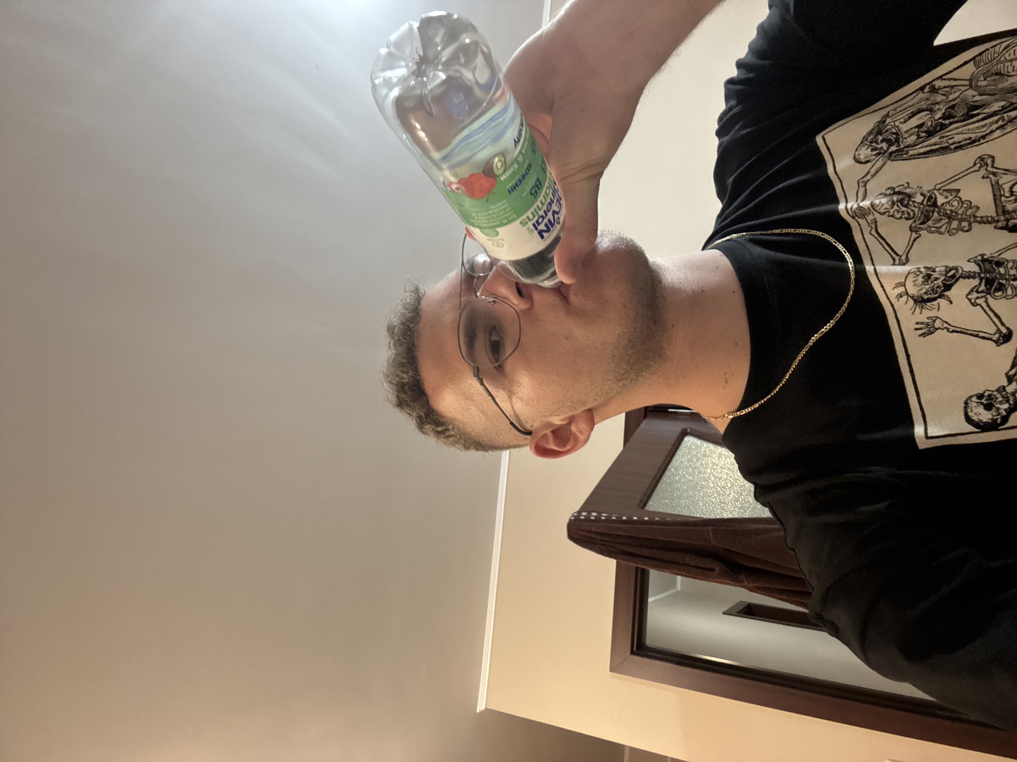 A man is depicted from the chest up, seen from a low angle, drinking from a plastic water bottle. He has short, dark, curly hair, stubble, and wears wire-rimmed glasses. A thin gold chain hangs around his neck, resting on a black t-shirt that features a large, off-white graphic print of various skeletal or anatomical figures.
The man is actively drinking, with the clear plastic bottle held to his lips. The bottle's label is light blue and green, displaying text such as "mineralins," "B5," and partially visible Cyrillic text including "АКТИВЕН" (Active) and "КОФЕИН" (Caffeine). Illustrations of a red berry and a green fruit are also visible on the label.
The background is simple, with a plain, light-colored wall or ceiling dominating the upper and left parts of the frame. To the right, a dark wooden door frame with a frosted glass panel is visible, suggesting an indoor setting. The image appears to be a candid moment, with no discernible emotion other than the act of drinking. No specific time of day or weather can be determined. The implied location context is Plovdiv, Bulgaria.