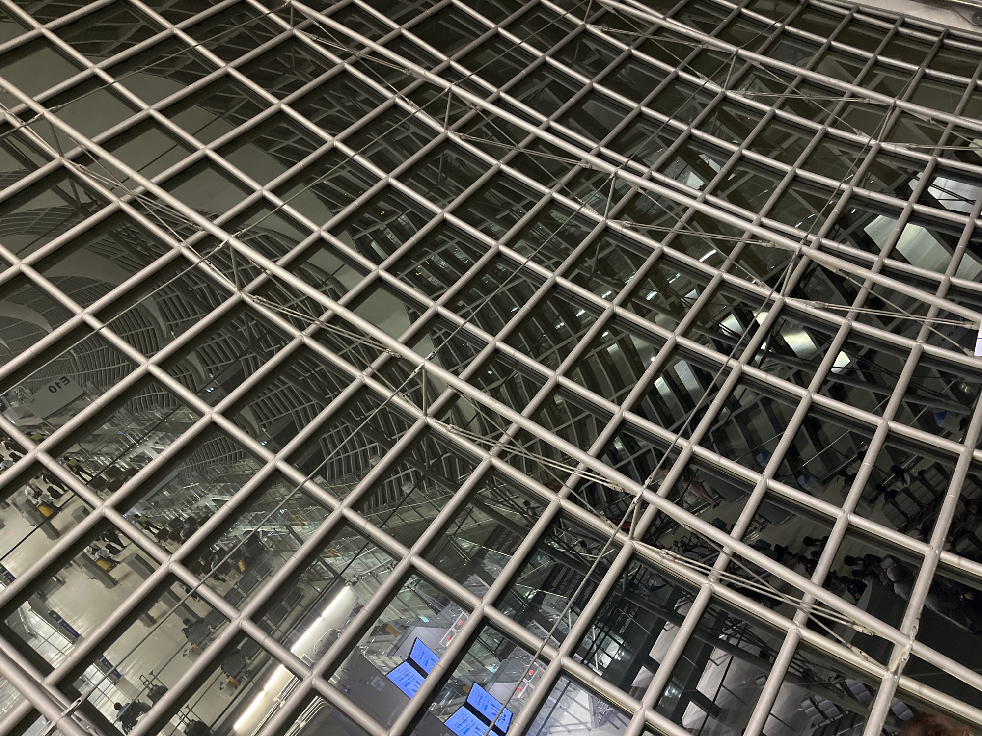 The image shows an interior view of a modern building, likely an airport terminal or a large public transport hub, with a distinctive lattice-like structure of metal beams and glass panels overhead. The perspective is looking upwards and diagonally through this structure.
Within the reflections and through the glass, several people can be seen. Many appear to be waiting, some with luggage, suggesting travel. There are also rows of seating visible in some areas. Digital displays or screens are present, one showing text that appears to be in English. The architectural style is sleek and contemporary, with a focus on open space and glass. The overall impression is one of a busy, well-lit, and modern interior.
The prompt requests identification of the location in Nong Prue Subdistrict, Thailand. However, there is no visual information within the image itself that definitively places it in Nong Prue Subdistrict or Thailand. The architectural style is common in many international airports and modern buildings globally. Without external information or context, it is impossible to confirm the specific location.