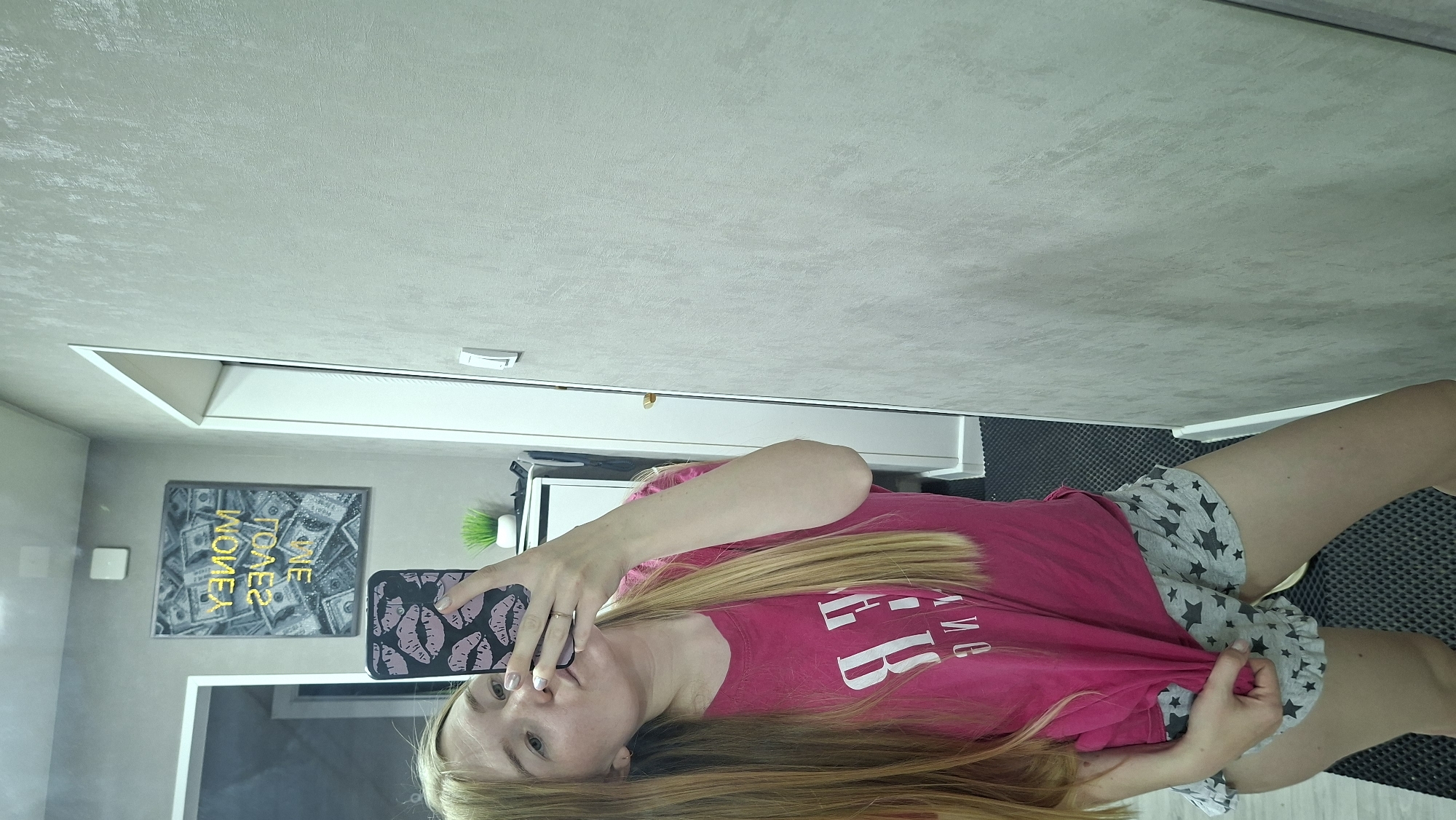 A woman is taking a selfie in an interior space. She has long blonde hair and is wearing a pink t-shirt and shorts with star patterns. She is holding a phone with a lip-print pattern case. Behind her is a wall with a picture of money and a white cabinet with a green plant. The exact location within the city of Chaikovsky, Russia, is not determinable from the provided image.