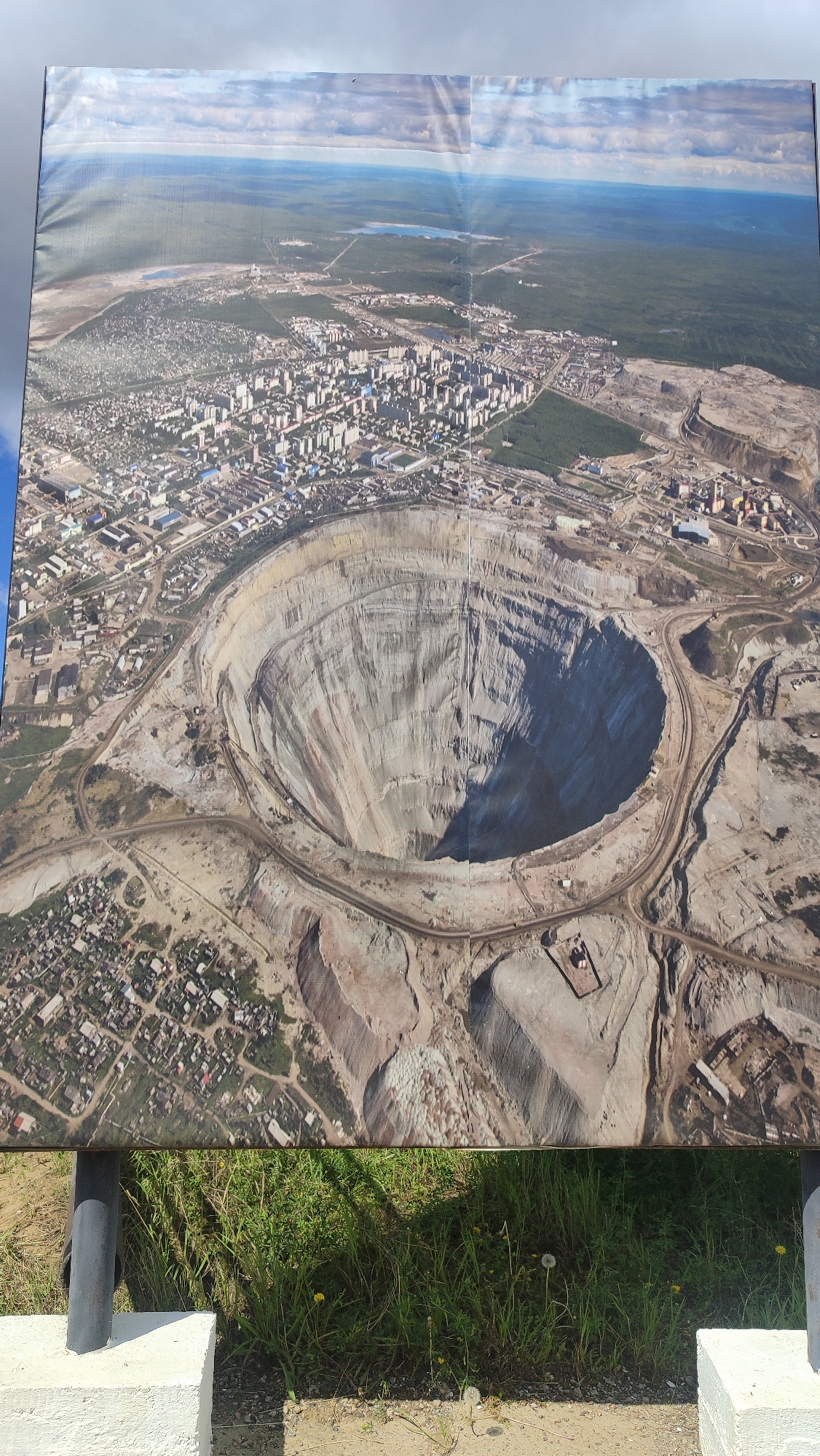 This is a photograph of a large, printed panorama displayed outdoors, likely at an informational exhibit. The panorama depicts an aerial view of the city of Mirny, Russia, with a prominent feature being a massive, deep open-pit diamond mine. The mine is circular with steep, terraced sides and a dark interior, suggesting significant depth. Surrounding the mine is the urban landscape of Mirny, characterized by densely packed apartment buildings and smaller residential areas. Beyond the city, the panorama shows a vast expanse of green forest and a body of water in the distance. The printed panorama is supported by a metal pole and is positioned in front of a grassy area with some yellow wildflowers and dandelions. The ground in front of the display consists of dirt and gravel. Two white concrete blocks are visible at the bottom corners, likely part of the display's foundation. The sky above is cloudy, with patches of blue visible.