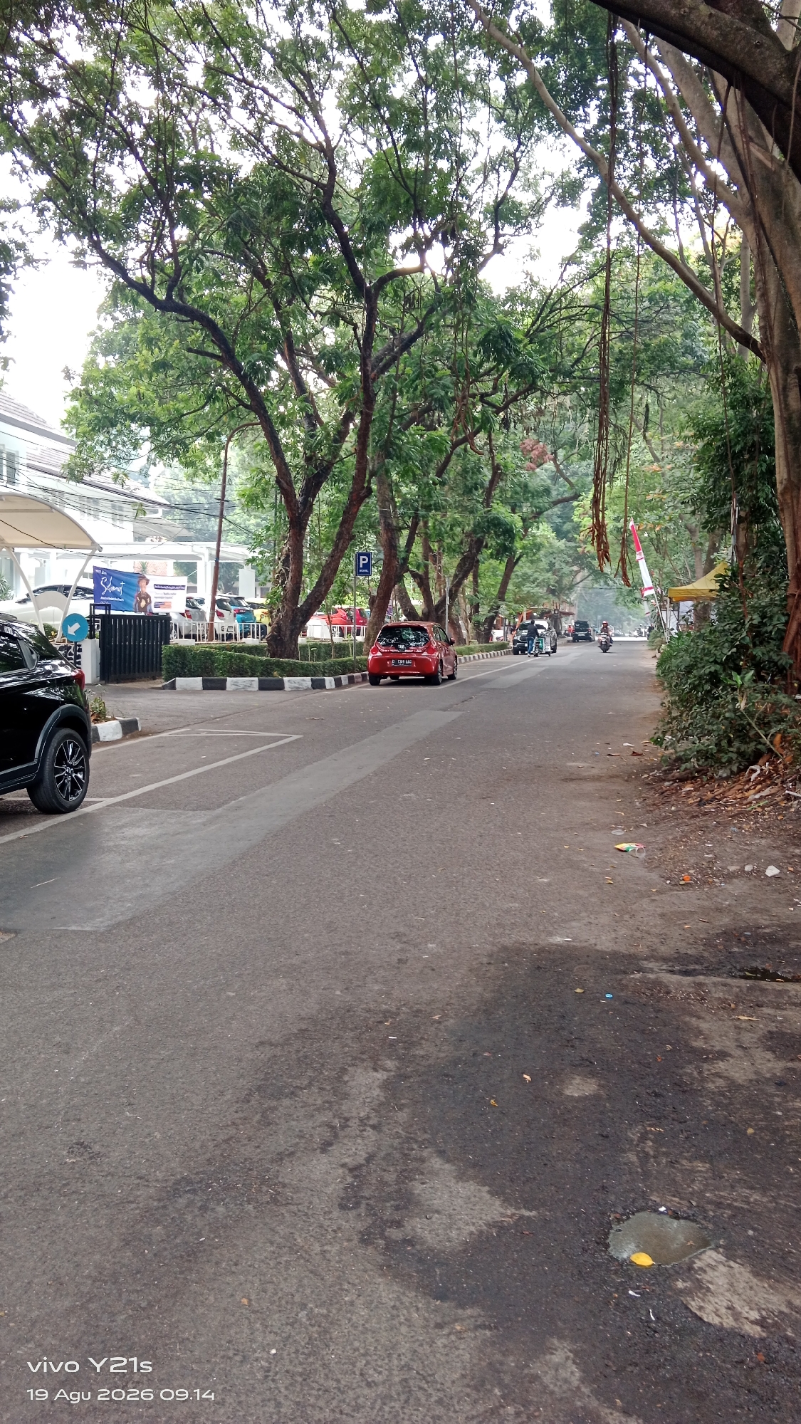 The image depicts a tree-lined street in Bandung City, Indonesia. A black SUV is parked on the left side of the road, partially in frame. A red hatchback is driving down the road, followed by a motorcycle. The street is paved and shows signs of wear, with some puddles and debris visible. Tall trees with dense green foliage overhang the road, creating a canopy effect. In the background, there are buildings and a parking lot with several cars. A blue parking sign is visible on the right side of the road. The sky is overcast. The timestamp "vivo Y21s 19 Agu 2026 09.14" is visible at the bottom left of the image.
