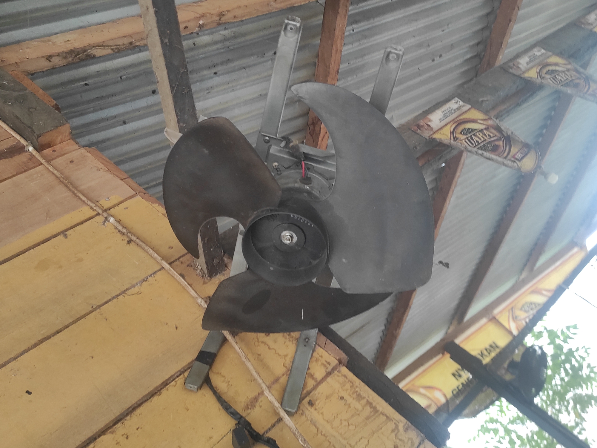 The image shows a ceiling fan with three dark-colored blades mounted on a wooden ceiling in a rustic, outdoor setting. The fan is attached to a metal bracket that is secured to wooden beams. The ceiling is made of yellow painted wooden planks, and the roof appears to be corrugated metal. There are some advertisements visible on the beams below the fan, one of which says "QUARA." The overall impression is of a simple, perhaps temporary, structure. The specific location in Lhokseumawe, Indonesia, cannot be determined from the image alone.