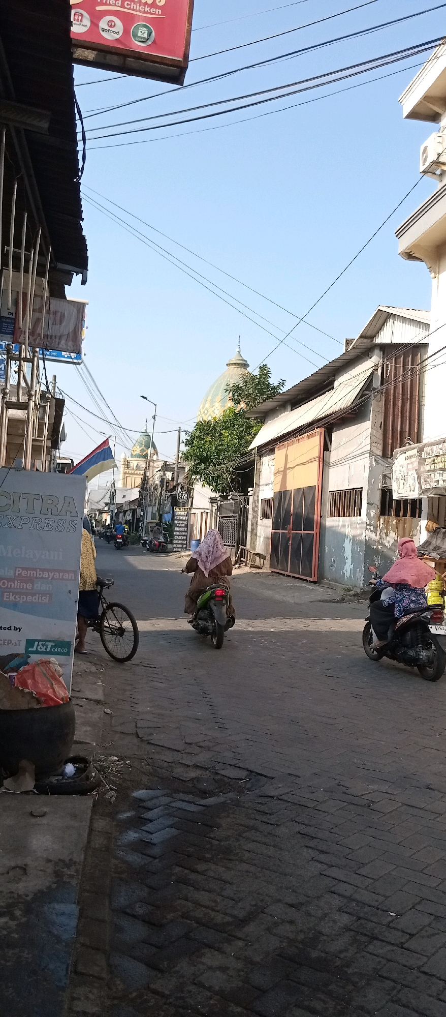 The image depicts a street scene in Wadungasri, Indonesia. In the foreground, a sign for "CITRA EXPRESS" is visible on the left, with a bicycle parked next to it. A woman wearing a brown dress and headscarf is riding a scooter away from the camera, heading down the street. Further down the street, another woman, also wearing a headscarf and a patterned blue shirt, is riding a scooter towards the camera. The street is paved with brick. Buildings line both sides of the street, with one building on the right having a corrugated metal roof and large orange doors. In the background, two domes, one larger and green with gold accents, and another smaller and green, are visible, suggesting the presence of a mosque or similar religious structure. Power lines crisscross the sky, and trees are present. The sky is clear and blue. The overall impression is of a typical street in a residential or commercial area.