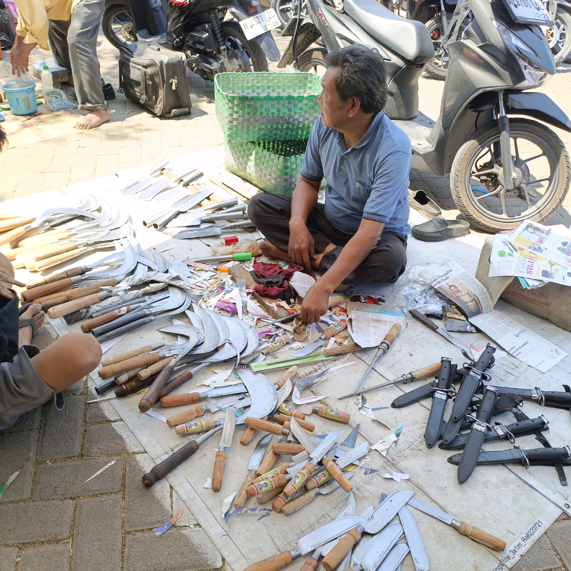 A man sits on the ground in Ambarawa, Indonesia, surrounded by numerous knives and sickles. He is wearing a blue polo shirt and dark pants. The ground is paved with bricks and covered in a white tarp, on which the tools are displayed. Various newspapers and scraps of paper are scattered among the items. In the background, motorcycles are parked, and a green woven basket is visible. A blue bucket and a bottle of liquid are also present on the ground.