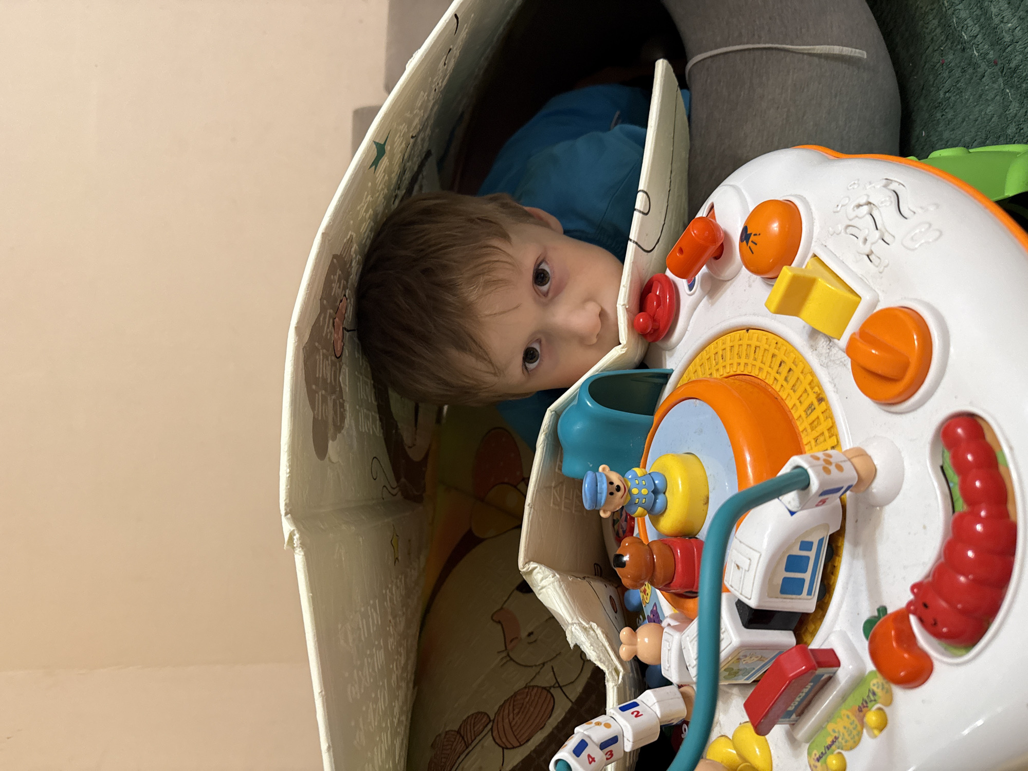 A young child is inside a cardboard structure, possibly a makeshift fort or play area, with a colorful toy activity center positioned near the opening. The child is wearing a blue shirt and has light brown hair. The toy activity center is white and orange, with various interactive elements such as buttons, knobs, and movable figures. Parts of the child's legs and gray sweatpants are visible behind the cardboard structure and activity center. The background is a plain wall, possibly inside a home. The location is not specified.