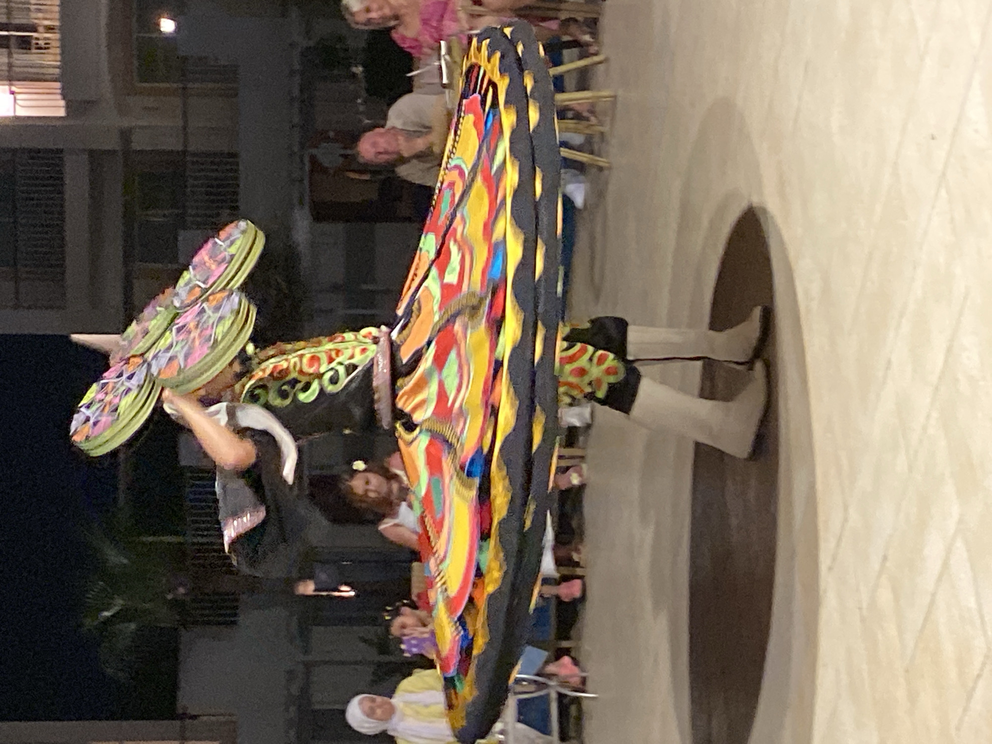 A male performer in a tanoun costume spins in a circle at night, creating a blur of color with his wide, layered skirt and headpiece. He wears white boots. In the background, several people are seated at tables, some watching the performance. A woman in a yellow headscarf and a child sit at a table on the left. A man and a woman are seated at a table on the right. The setting appears to be an outdoor area with a tiled floor and buildings in the background, possibly in Sharm El Sheikh, Egypt, given the presence of a tanoun performer. Palm trees are visible in the dark background.
