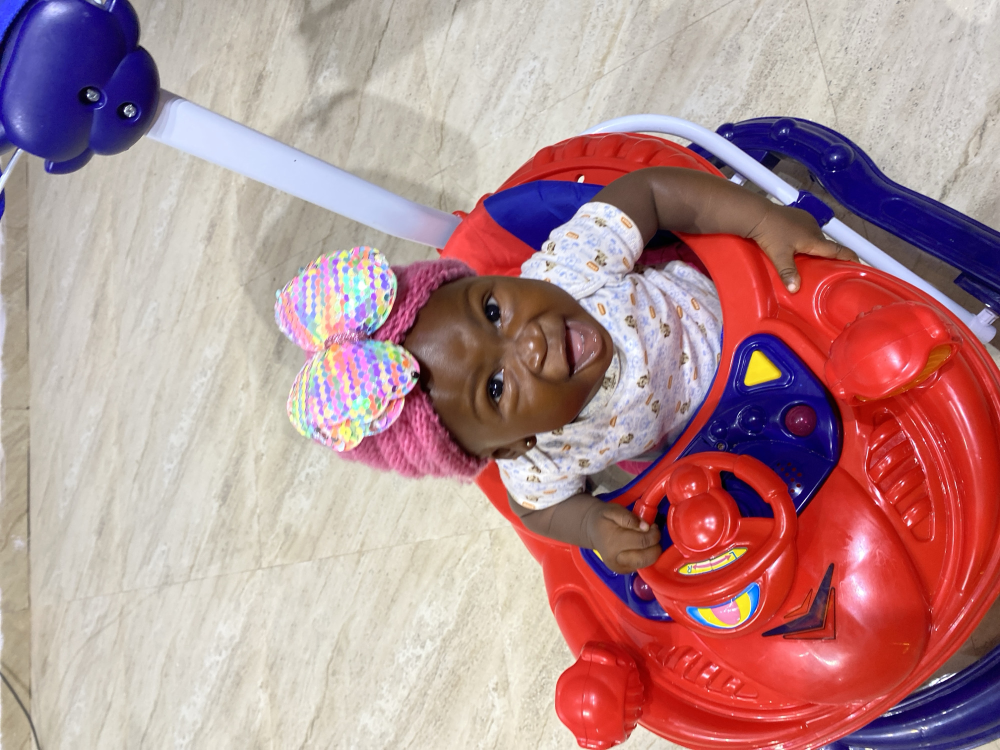 An infant girl wearing a pink knitted cap with a rainbow sequined bow sits in a red and blue plastic baby walker. The baby is looking at the camera with a wide smile and her mouth open. The walker has a steering wheel and several colorful buttons. The background is a beige tiled floor. The image appears to have been taken indoors, likely in a residential setting in Gura Top, Nigeria.