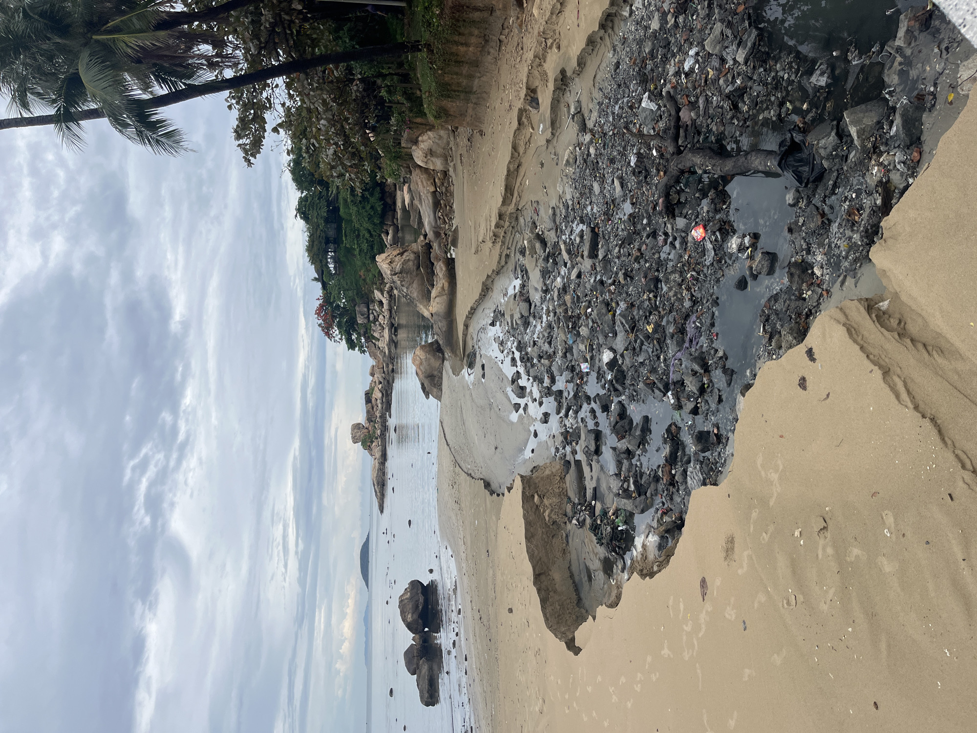 The image depicts a heavily polluted beach, possibly in or near Bac Nha Trang Ward, Vietnam. The foreground shows a sandy beach area with footprints, leading to a sharp drop-off into a channel filled with dark, stagnant water and garbage. The channel is choked with plastic debris, rocks, and organic waste. In the background, there is the ocean, with rock formations and a cloudy sky above. The shoreline is lined with trees and vegetation.