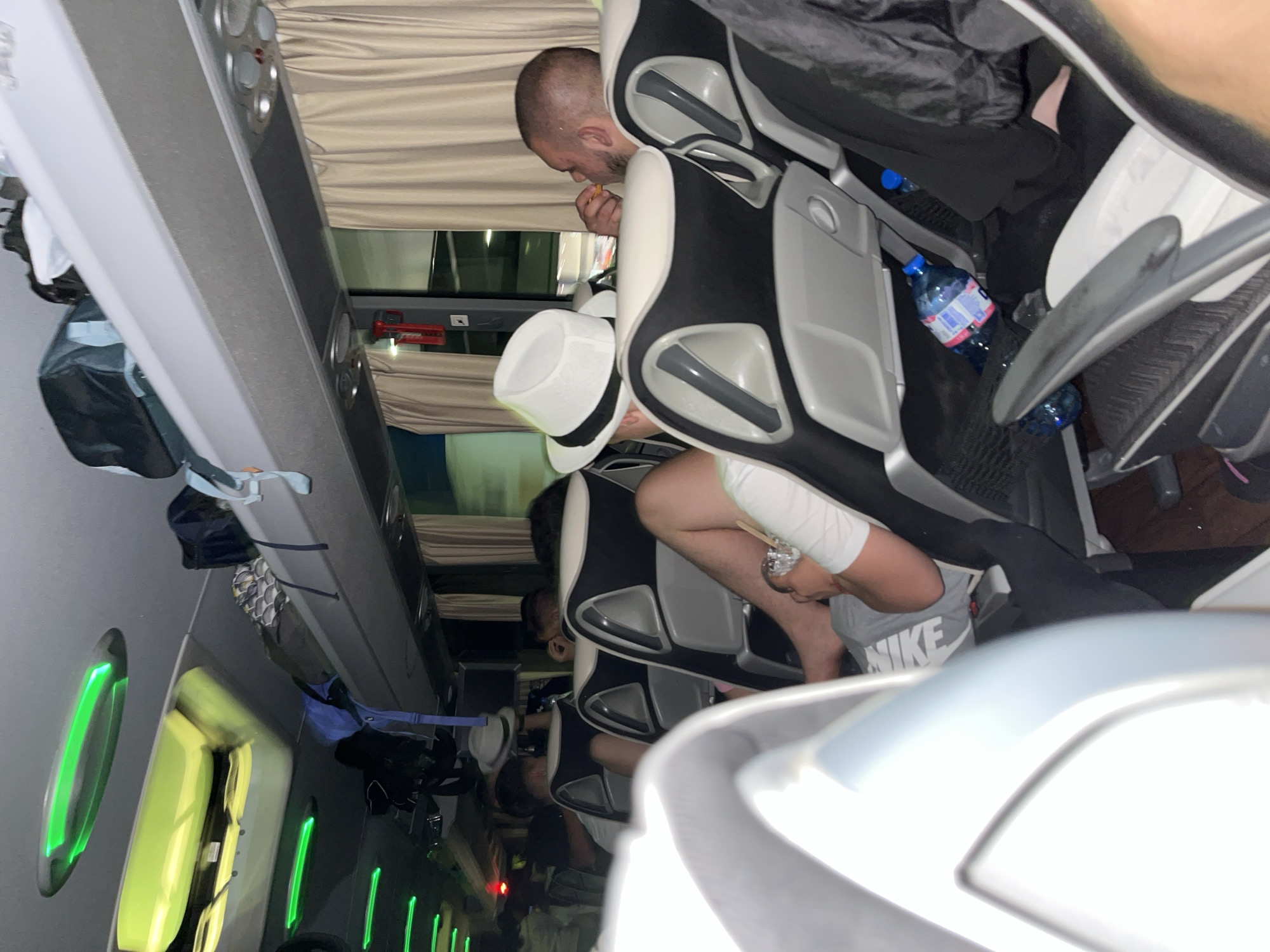 The scene appears to be inside a passenger bus or shuttle, possibly in transit to Popovitsa, Bulgaria, although this is not definitively confirmed by the image. Several people are seated in reclining passenger seats. One man is eating, and another wears a white fedora and grey Nike shorts. There are water bottles visible. The interior of the bus has overhead compartments. The windows are visible on the left side of the frame and have an emergency exit sign. Green lighting is present along the bus interior.