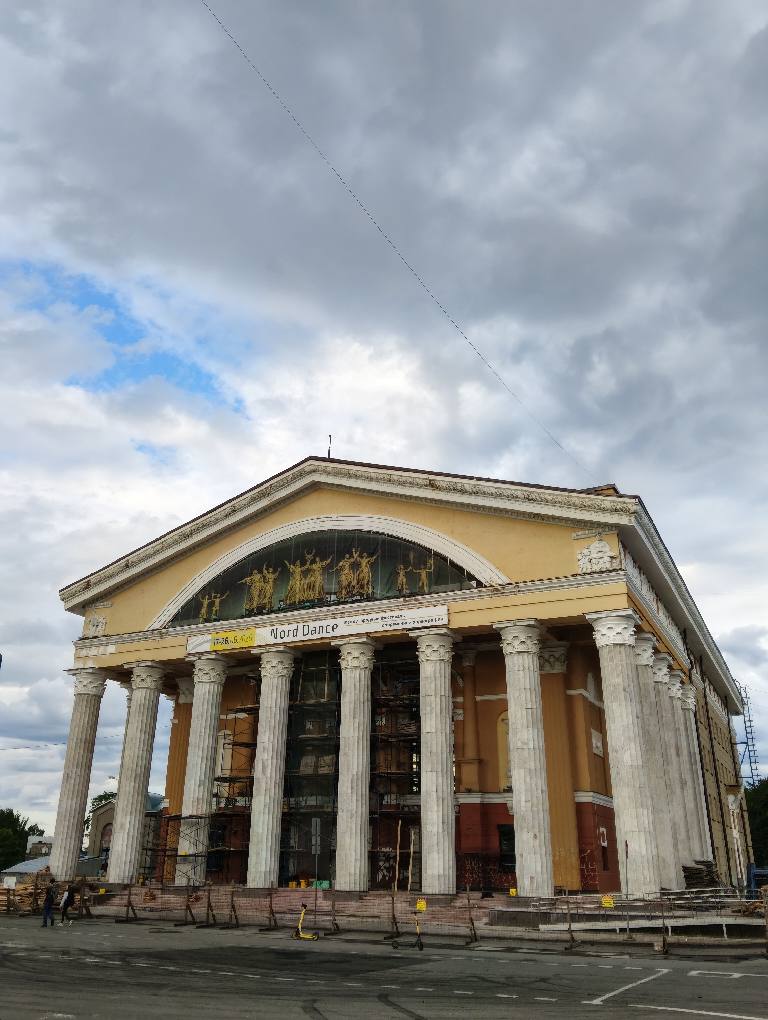 The image shows the National Theatre of the Republic of Karelia in Petrozavodsk, Russia. The building is a grand structure with a yellow facade, white columns, and an arched window decorated with golden figures. A banner hangs across the facade, announcing "Nord Dance" and a date range of 17-26.06.2025. The building is partially covered in scaffolding, suggesting ongoing renovation or construction work. In front of the building, there are several yellow electric scooters parked on the sidewalk, and two people are walking on the pavement. The sky is overcast with dramatic clouds.