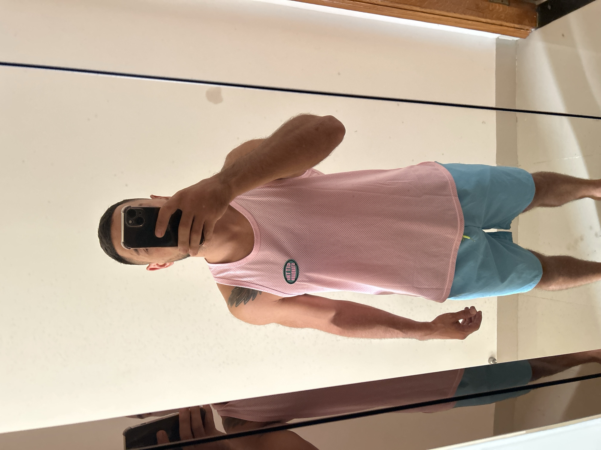 A man takes a mirror selfie, wearing a pink mesh tank top with a circular logo on the left chest and blue swim trunks. He has short dark hair and a beard. His left arm has a tattoo. The mirror is positioned in a room with light-colored walls. A sliver of a wooden doorframe is visible on the right.