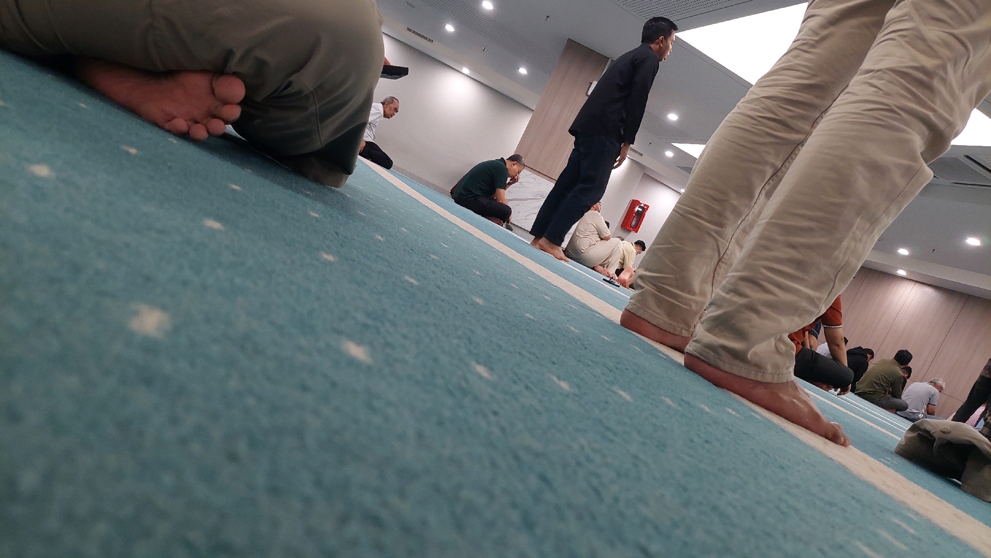 The image captures an indoor scene, likely a mosque or prayer hall, with a distinctive turquoise carpet featuring a white stripe and scattered white dots. The low-angle perspective places the viewer close to the floor, emphasizing the texture of the carpet and the lower halves of several individuals. In the foreground, the bare feet and lower legs of two people are visible, one wearing light-colored, wrinkled trousers, and the other wearing darker trousers.
Further into the scene, a group of men are seated or in prayer positions on the carpet. One man in a black shirt and dark pants is standing, appearing to lead or observe. Other men are dressed in various casual attire, some in green shirts, and one in a light-colored outfit. Their postures suggest they are engaged in prayer.
The environment is brightly lit with overhead fluorescent lights, and a modern ceiling with visible tracks is present. A red fire extinguisher is mounted on a light-colored wall in the background, and a wooden-paneled wall is also visible. The overall impression is one of a communal prayer gathering within a clean, well-lit indoor space. The presence of men in prayer suggests a religious context, and given the common architectural and carpeting styles, it is plausible this scene is located in a mosque in Bekasi, Indonesia, a city known for its significant Muslim population and numerous places of worship.