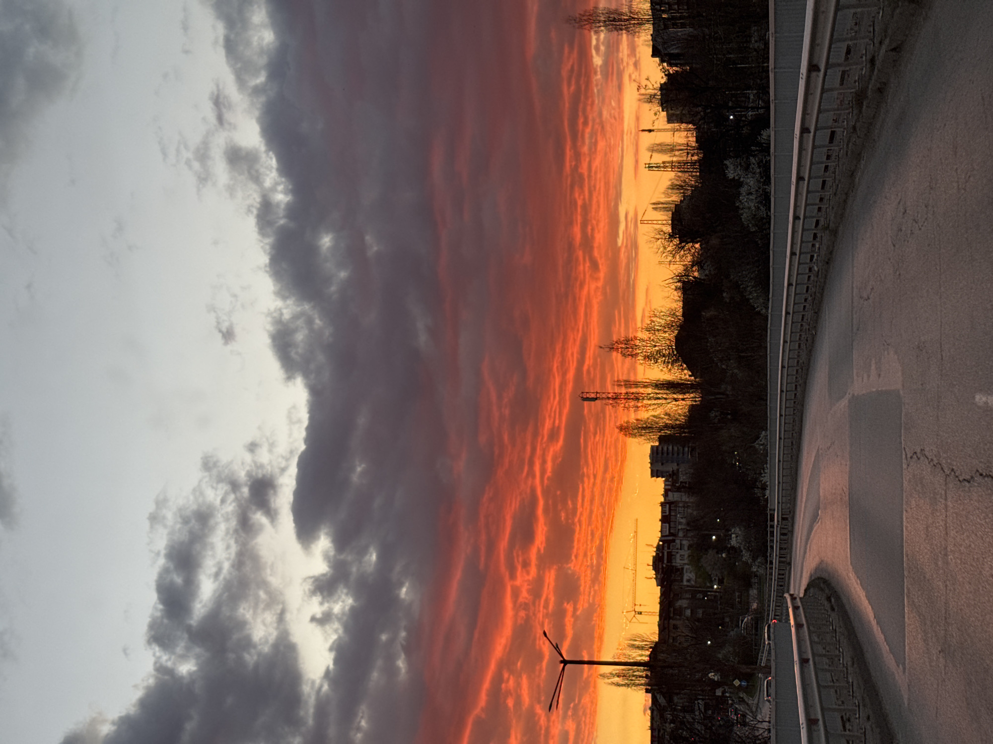 The image captures a dusk scene in Sofia, Bulgaria. The foreground is a road surface, curving gently to the right, with a metal guardrail running alongside it. The road appears to be slightly wet, reflecting ambient light. Beyond the guardrail, there is a row of bare trees and buildings silhouetted against a vibrant, orange-red sky. In the middle distance, a construction crane is visible, suggesting urban development. The sky itself is a dramatic mix of grey clouds on the left, transitioning into intense orange and red hues towards the horizon, indicating sunset or sunrise. The overall scene conveys an urban environment transitioning from day to night under a striking sky.