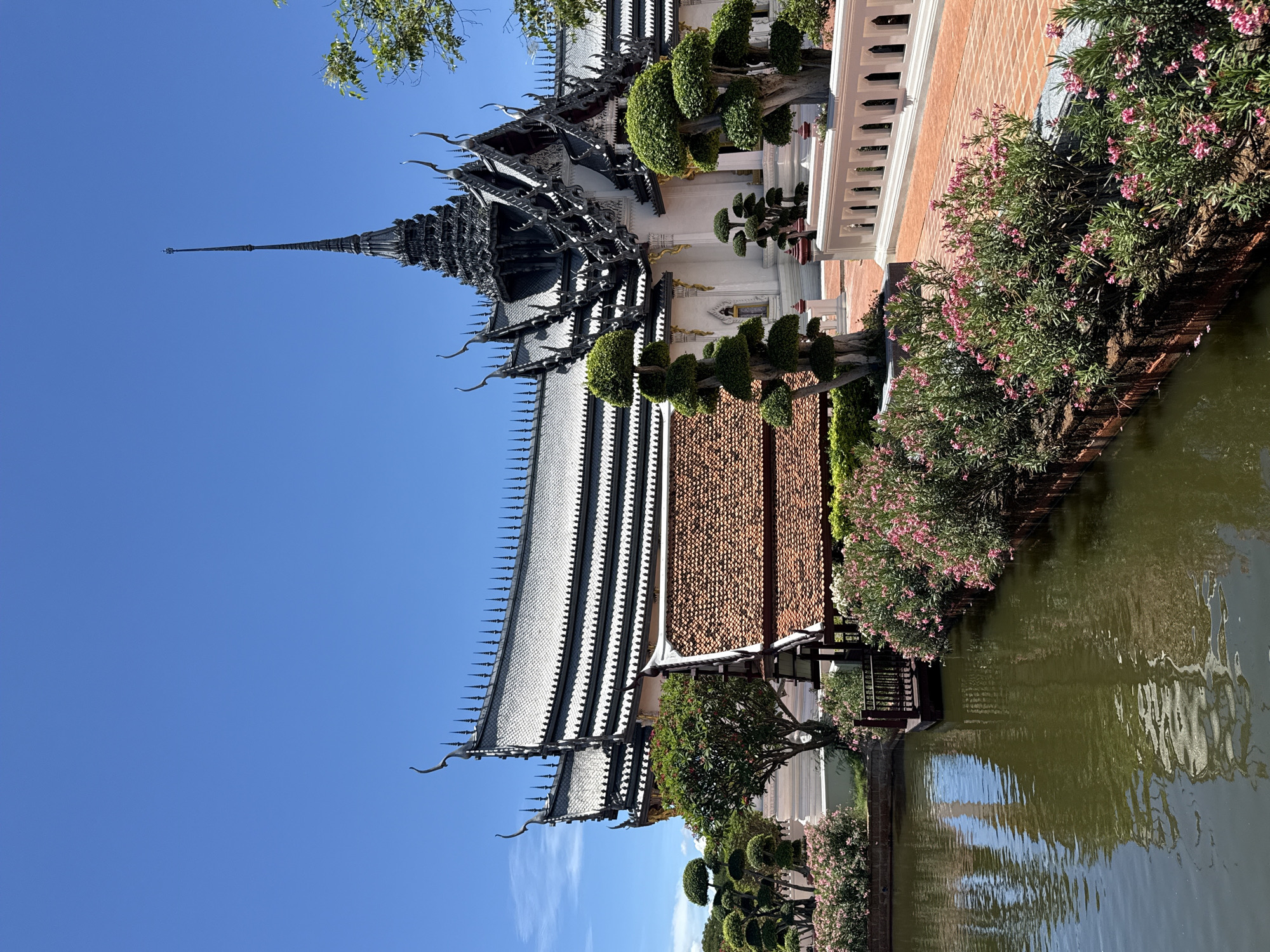 The image depicts a building with a tiered, ornamental roof structure in Bang Pu Subdistrict Municipality, Thailand. The building is adjacent to a body of water. The architectural style is characterized by intricate details and multiple roof levels. The lower part of the building is white with a red brick border. Mature trees and flowering plants border the body of water. The sky is clear blue.