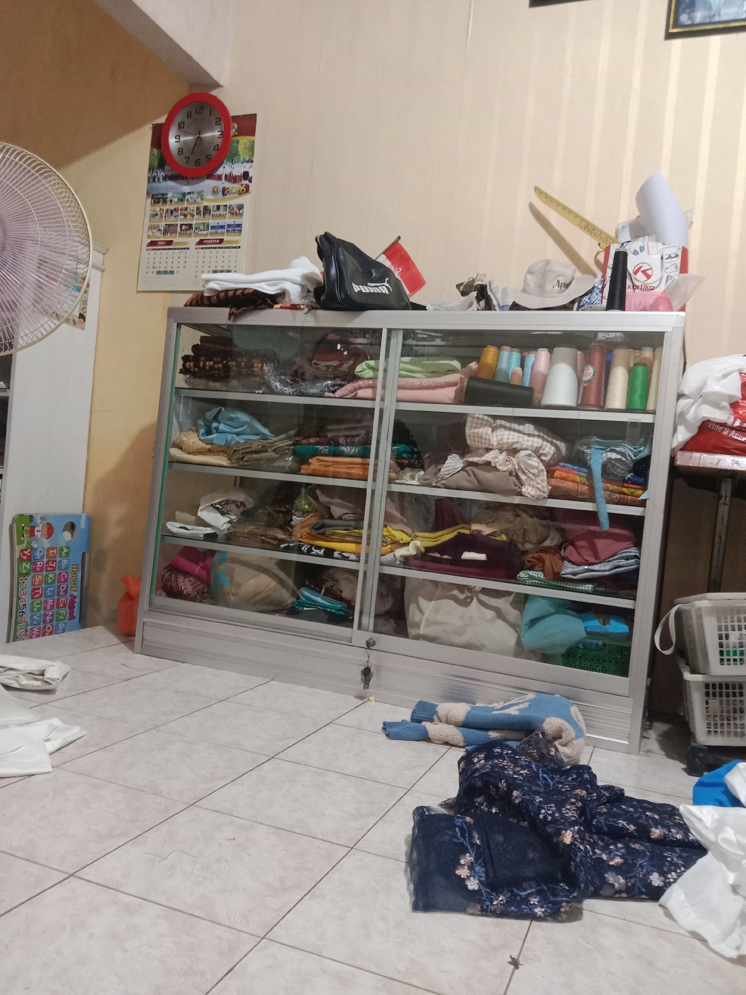 This image captures an indoor scene, likely a small shop or a room dedicated to tailoring or fabric sales, situated in Madiun, Indonesia. The focal point is a silver-framed glass display cabinet filled with various fabrics, threads, and possibly finished garments. The cabinet has multiple shelves, each neatly organized with folded textiles in different colors and patterns. To the left of the cabinet, a white oscillating fan is visible, and a colorful children's educational mat with alphabet and numbers is propped against a wall. Above the cabinet, a red-faced analog clock is mounted on the wall, accompanied by a calendar showing the months of July and August, with the year "2006" clearly visible, suggesting the photo was taken in that year. On the top shelf of the cabinet and on the wall above it, various items are placed, including bags, hats, spools of thread in multiple colors, and other miscellaneous accessories. The floor is tiled in a light beige or gray color. In the foreground, several pieces of dark blue and floral patterned fabric are laid out on the floor, along with some white fabric to the left. To the right of the cabinet, a white laundry basket is visible, partially obscured. The lighting appears to be a mix of ambient indoor light and potentially artificial light, casting soft shadows. The overall impression is one of a working space with an organized yet slightly cluttered atmosphere.