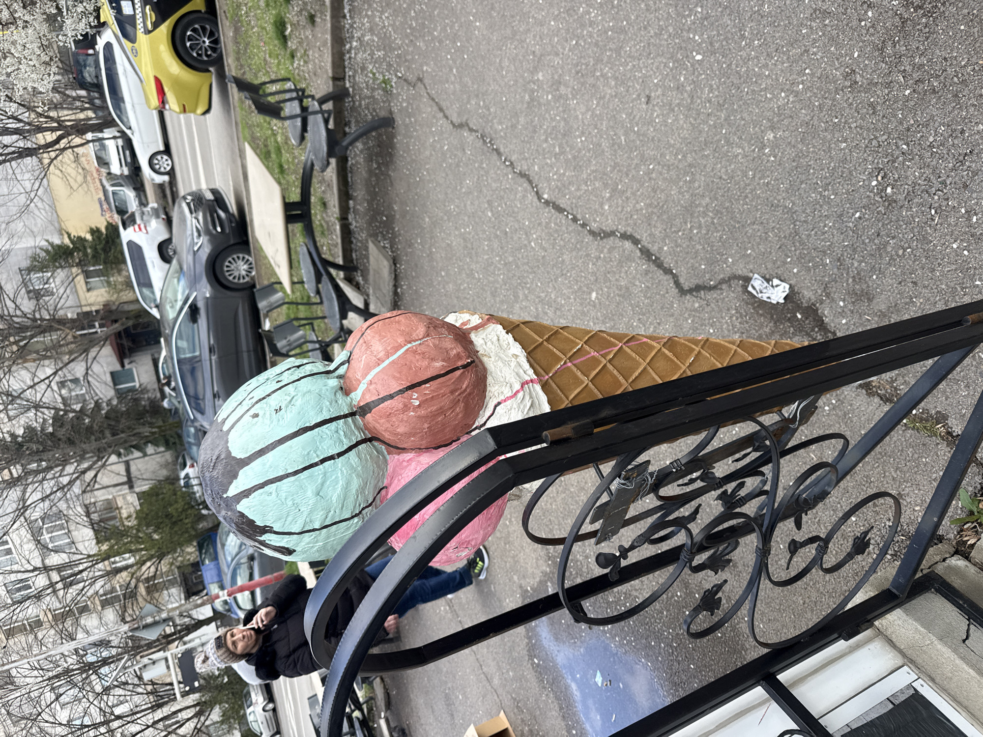 Outside a building in Sofia, Bulgaria, a very large fake ice cream cone is mounted on a wrought iron decorative stand. The ice cream cone has three scoops: mint chip, pink, and brownish-red. A woman in a black jacket and gray knit hat is standing to the left of the ice cream cone. Cars are parked on the street behind the ice cream cone, and a few park benches are nearby. The ground is concrete.