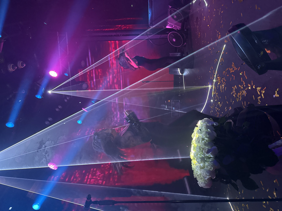 A stage is illuminated with blue and pink spotlights and laser beams, revealing two figures. One figure is a woman with long hair and black clothing, possibly performing or posing amidst the light show. A bouquet of white roses sits prominently in the foreground. Golden confetti litters the stage. Other stage elements visible include black equipment and a backdrop with red and white designs. The image has a slightly tilted perspective.