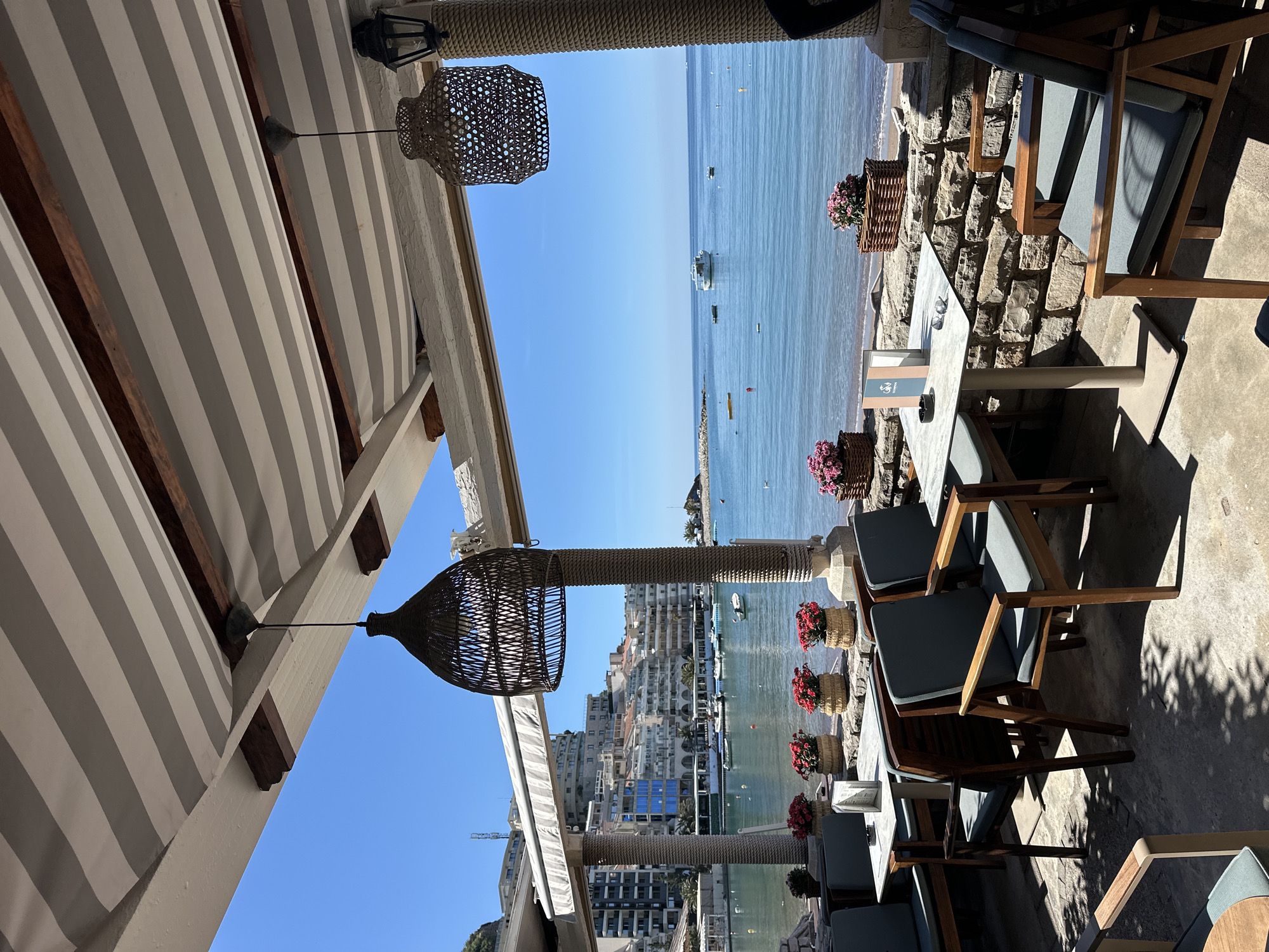 The image depicts an outdoor dining area in Boreti, Montenegro, overlooking the sea. The seating area comprises wooden-framed chairs with gray cushions arranged around square tables. Wicker baskets filled with pink flowers line the stone wall separating the dining area from the beach. Overhead, a striped awning provides shade, with two wicker pendant lights hanging from the structure. Boats are visible on the water. Buildings are situated along the coast in the background. The sky is clear.