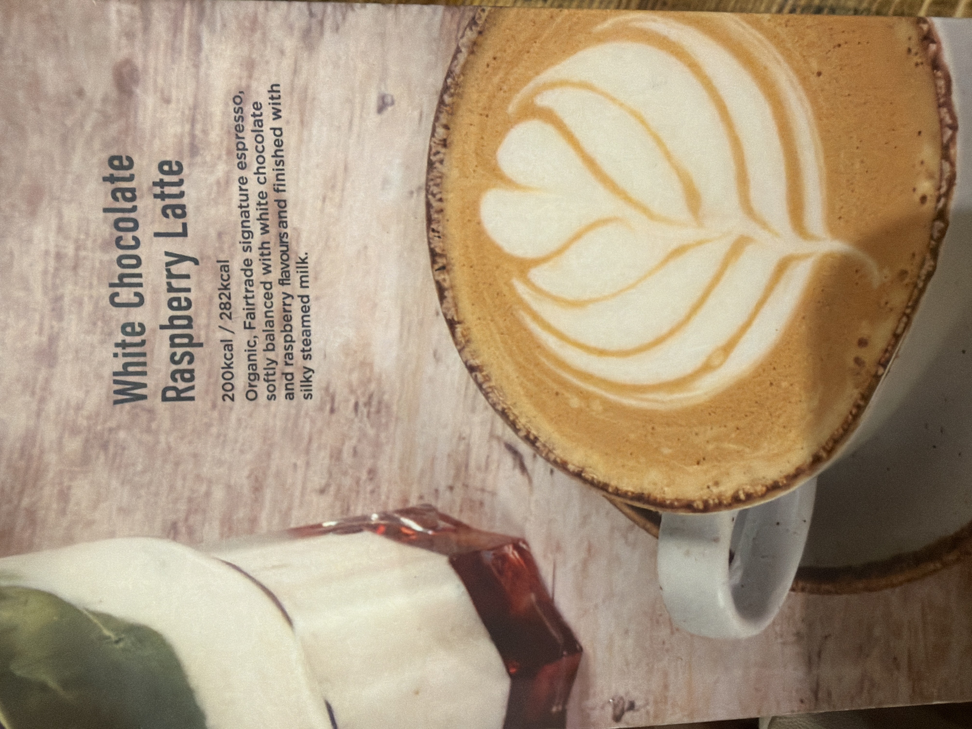 The image shows a promotional shot of a "White Chocolate Raspberry Latte." A frothy, latte art-adorned latte is the main subject of the image, presented in a mug, sitting on a saucer. To the left of the mug is a partially visible glass, likely containing ingredients related to the latte. Text to the left of the drinks advertises the beverage, noting its calorie count, ingredients (including organic, Fairtrade espresso, white chocolate, and raspberry flavors), and texture (silky steamed milk). The scene appears to be set in a cafe or restaurant, potentially in Mole Valley, United Kingdom, given the context.