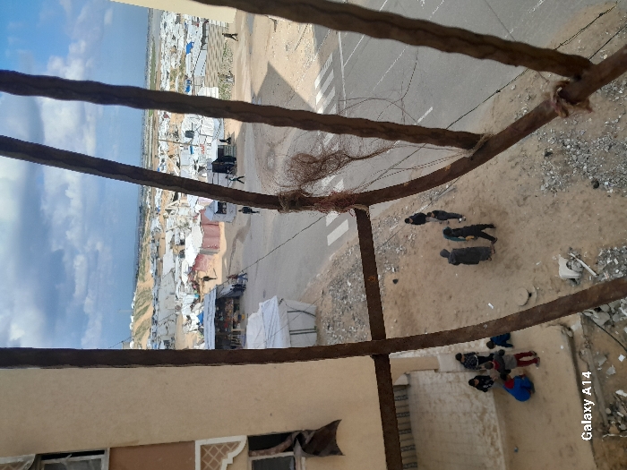 The image presents an elevated, downward-angled view of a displacement camp in Khan Yunis, Palestinian Territory. The scene is framed in the foreground by prominent rusty, dark brown metal bars, with reddish-brown fibrous material intertwined around a vertical bar on the right. To the lower left, a beige multi-story building is visible, featuring windows, one of which has a dark fabric hanging outward.
On the ground directly beneath the viewpoint, a patch of dirt and scattered rubble accommodates two groups of people. On the left, approximately 5-6 children, dressed in varied colored clothing including red and blue, are gathered on a concrete slab near the building, appearing to interact on the ground. To their right, 3-4 adults or older youths, predominantly in dark clothing, are standing or walking on the dirt.
A paved road with a white crosswalk horizontally traverses the midground, separating these individuals from a sprawling temporary settlement. This settlement consists of numerous shelters, primarily white tarpaulin tents, interspersed with more structured dwellings or market stalls constructed from materials such as corrugated metal, some painted in faded red hues. Several individuals are visible walking on the road and navigating the pathways within the dense camp.
The camp extends into the distant background, covering a vast area. Beyond the camp, sandy hills dominate the horizon under a partly cloudy sky. Faint structures, possibly wind turbines, are discernible atop the distant hills.