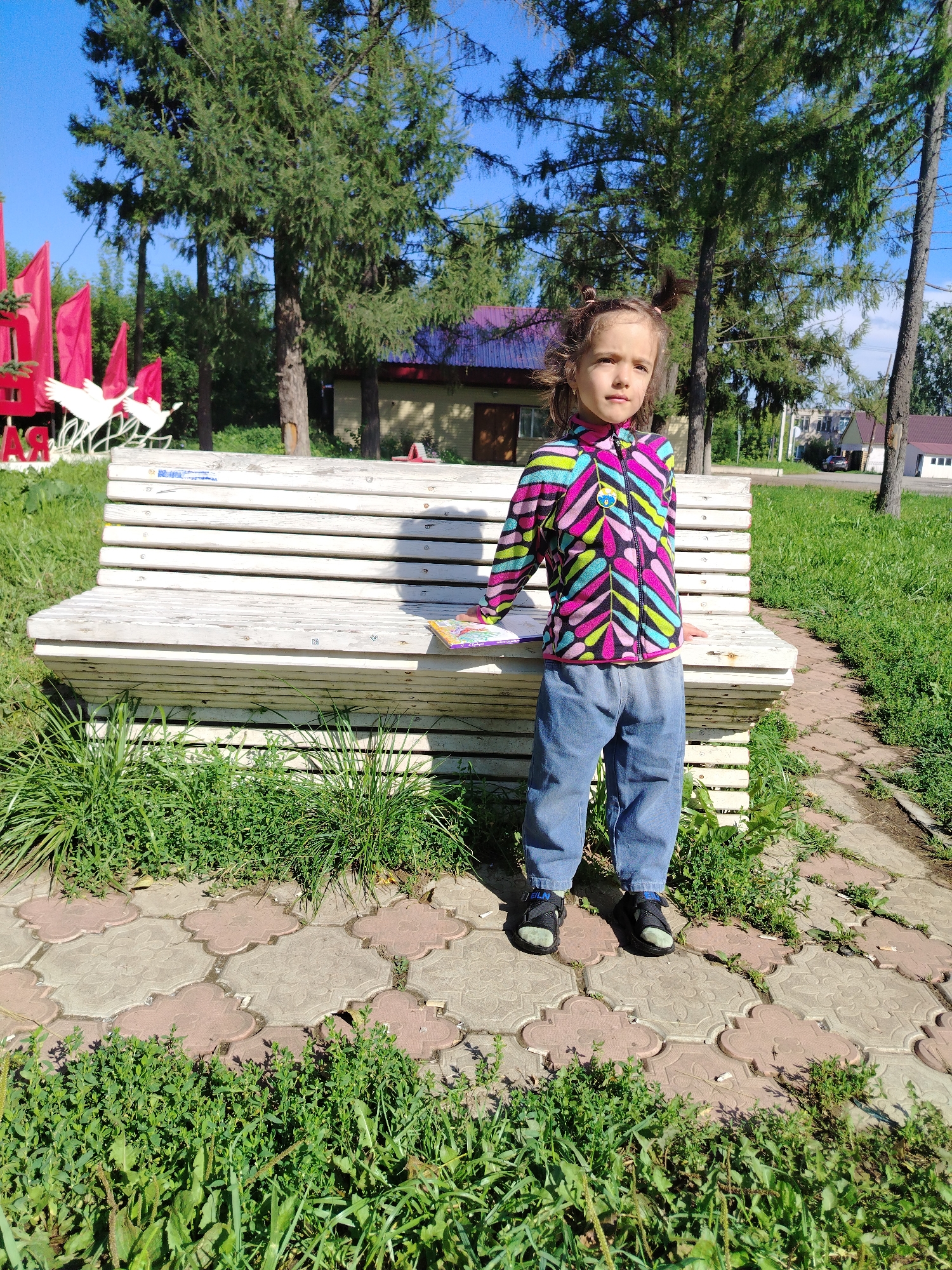 In Kuyeda, Russia, a young girl stands on a decorative paved path in a park. She is wearing a colorful jacket with a zigzag pattern in pink, green, blue, and black, and blue jeans. Her hair is styled in two pigtails. She is looking towards the right side of the frame. Behind her, a white wooden bench with horizontal slats is visible, with a book or magazine resting on its seat. The bench is surrounded by green grass and other vegetation. In the background, several tall trees are present, along with a building with a purple roof and red flags in the distance. The sky is clear and blue.