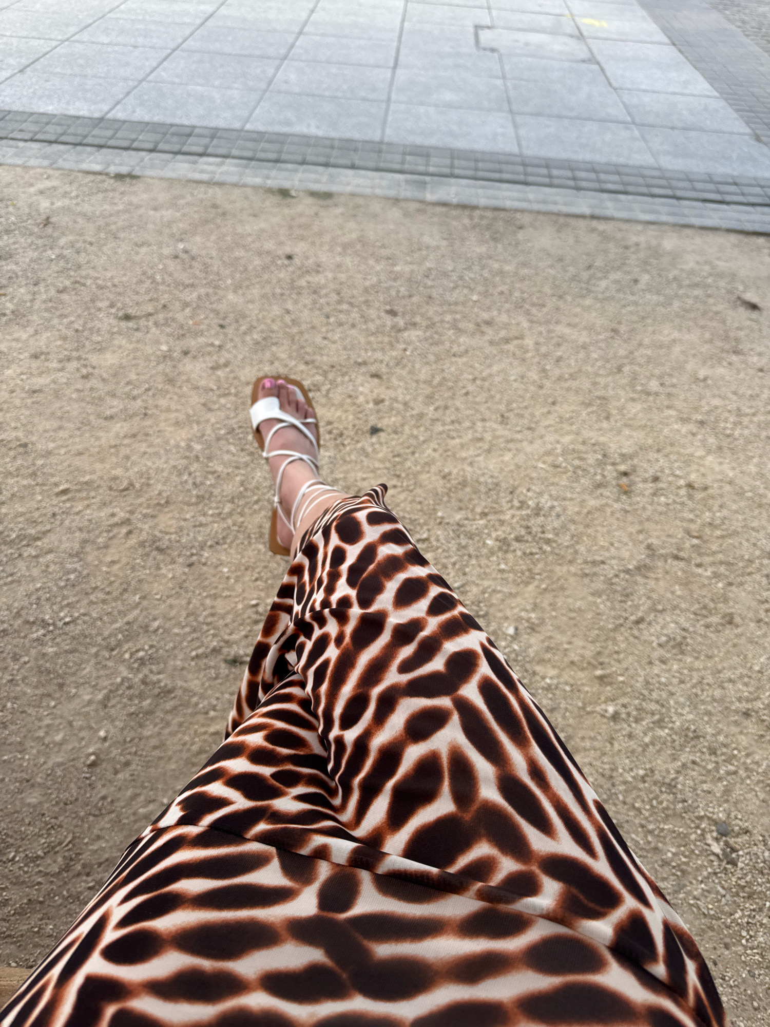 A person's legs are shown from the knees down, seated on the ground in a park or public space. The person is wearing a long, flowing skirt with a brown and white giraffe print pattern. On the feet are white strappy sandals, with one foot visible with painted pink toenails. The ground is covered with small gravel and sand. In the background, a section of paved walkway made of square grey tiles is visible. The scene appears to be outdoors.