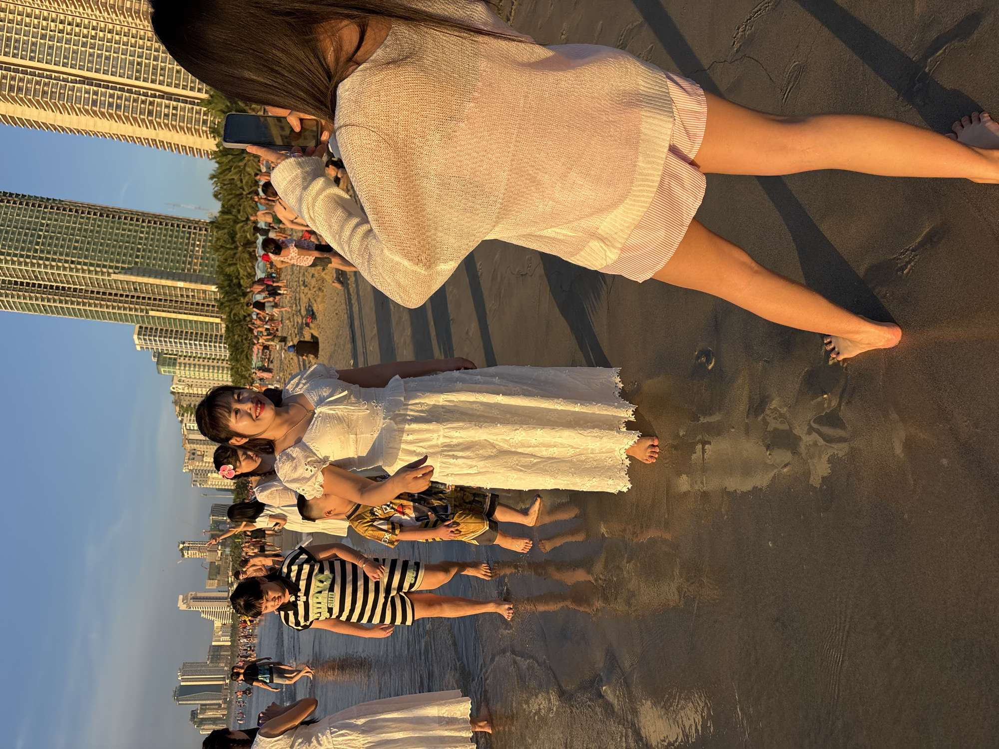 In Đà Nẵng, Vietnam, along the beach, a group of people are seen in shallow water with wet sand beneath their feet, reflecting the warm light of the setting sun. In the foreground, a woman is capturing a photo with her smartphone of another woman in a white dress. Behind them, several other individuals, including children, are walking along the shoreline. In the background, tall buildings line the coastline.