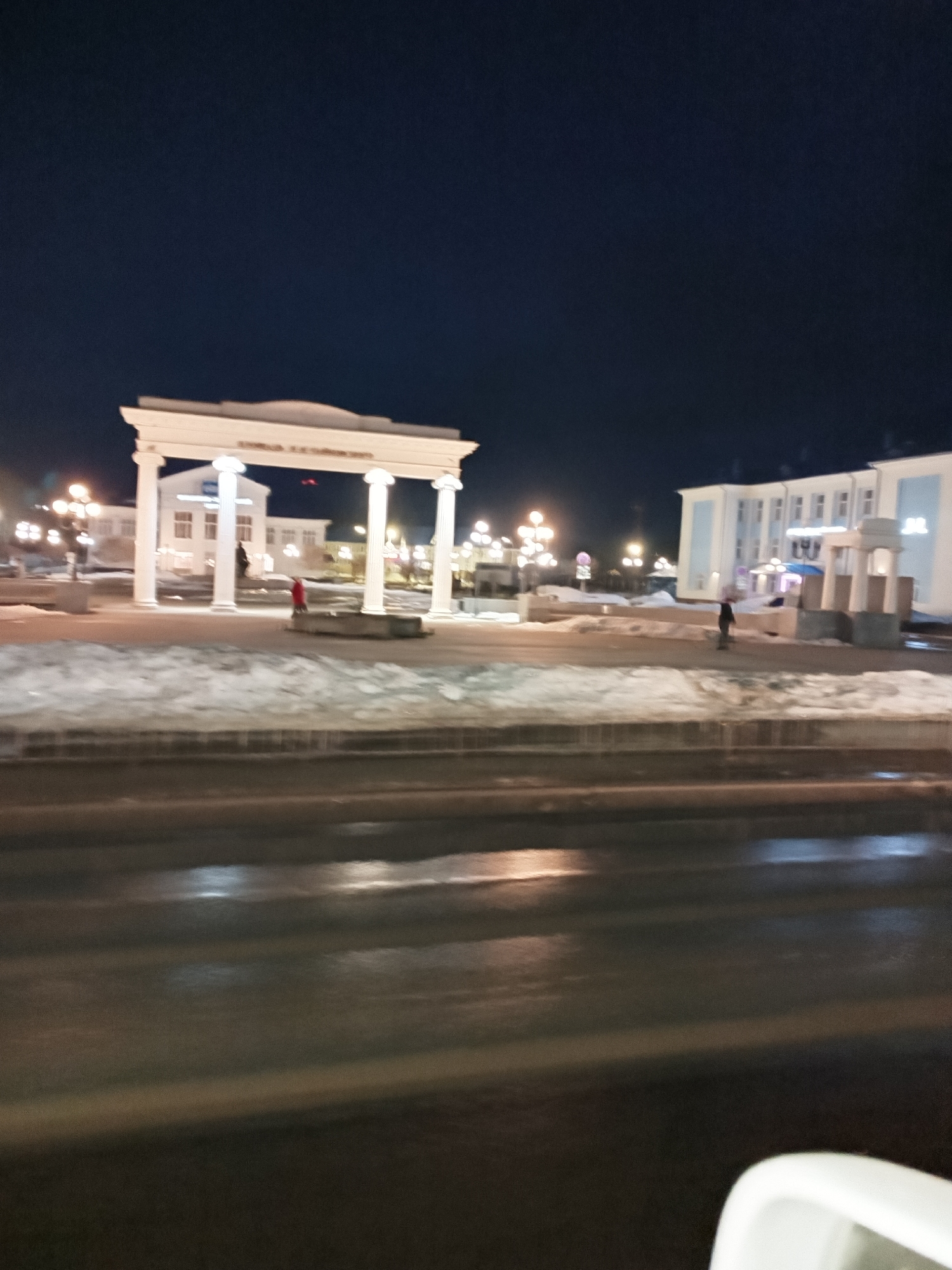 The image depicts a nighttime scene in Chaikovsky, Russia. In the background is the city's prominent white, colonnaded archway. A light dusting of snow covers the ground, interspersed with dark patches. The foreground reveals a wet road surface, with reflections of lights. A person in a dark coat is walking near the building to the right. Another person in red clothing is walking towards the archway. Streetlights illuminate the scene. A portion of the interior of a car is visible in the lower right corner.