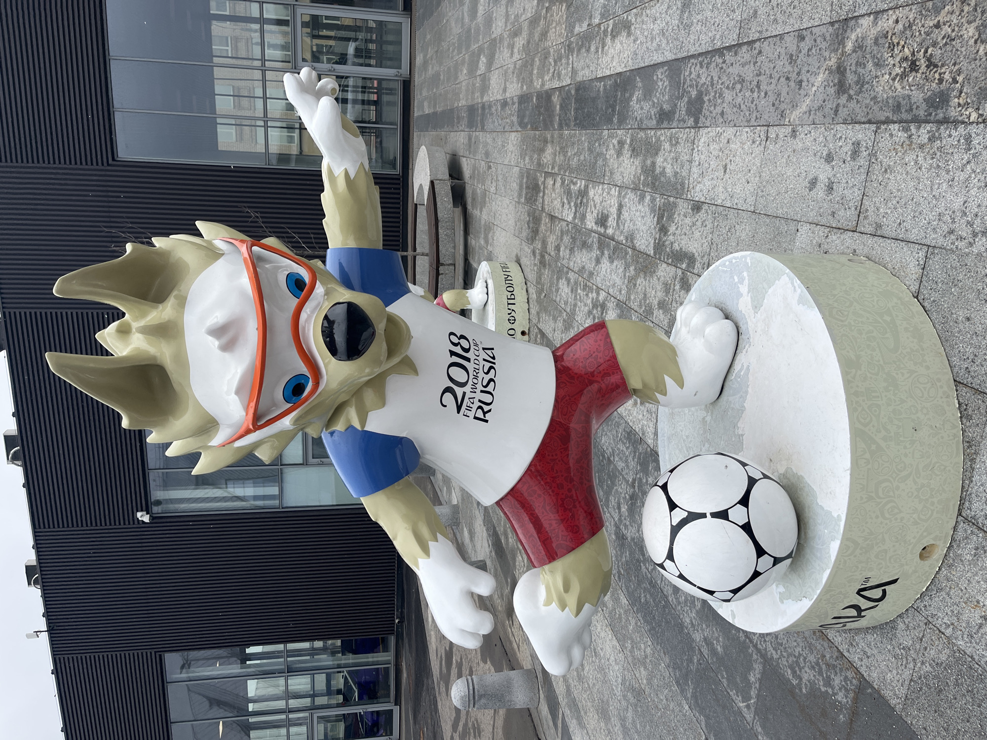 A statue of Zabivaka, the mascot for the 2018 FIFA World Cup, is positioned on a white, round base. Zabivaka is depicted as a wolf, wearing orange goggles and a white t-shirt with the "2018 FIFA World Cup RUSSIA" logo. The wolf's left arm is raised, and he is wearing red and white shorts. A soccer ball rests next to his foot on the base. The statue is located outdoors, on a gray tiled surface. A modern building with dark, horizontal lines and reflective windows is visible in the background. The location is Moscow, Russia.
