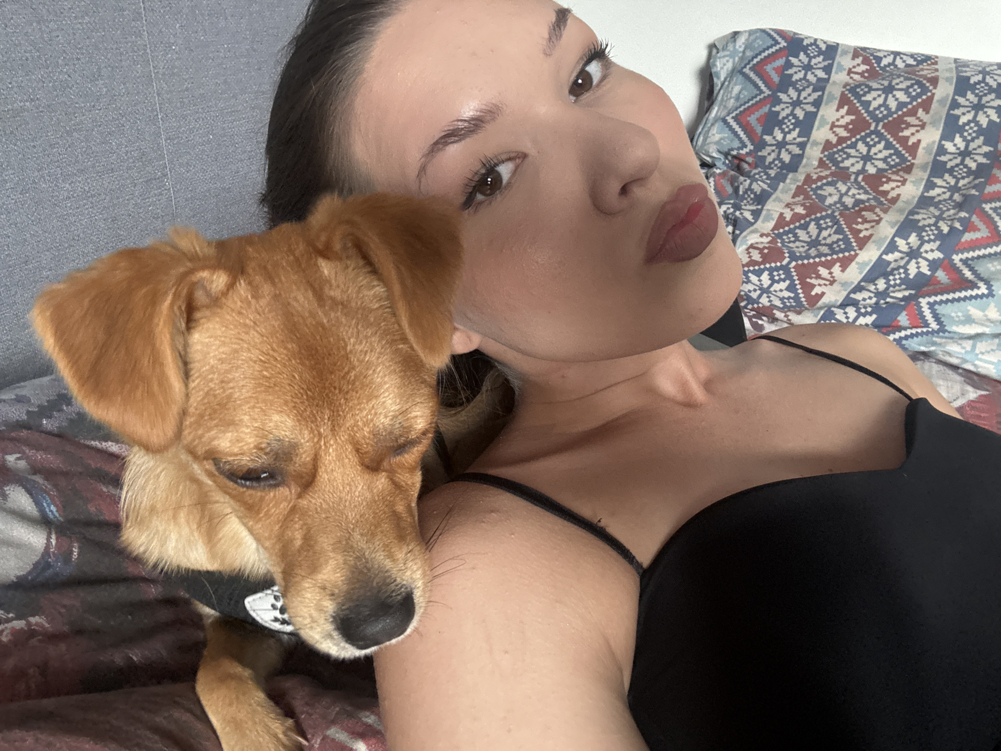 The image shows a woman and a small dog in a close-up shot, taken from a slightly high angle. The woman is lying down, with her head tilted and her eyes looking at the camera. She has dark hair, well-defined eyebrows, and light makeup. Her lips are slightly parted, and she is wearing a black tank top. The dog, a small breed with light brown fur, is nestled beside her, with its head resting near her face. The dog is wearing a black collar with a white emblem. They are on a surface that appears to be a bed, with a patterned pillow visible in the background. The background also includes a gray textured surface, possibly a headboard or wall, and a dark, patterned fabric underneath them. The lighting is soft, suggesting an indoor environment. The location in Vishnevka, Russia is not discernible from the image.
