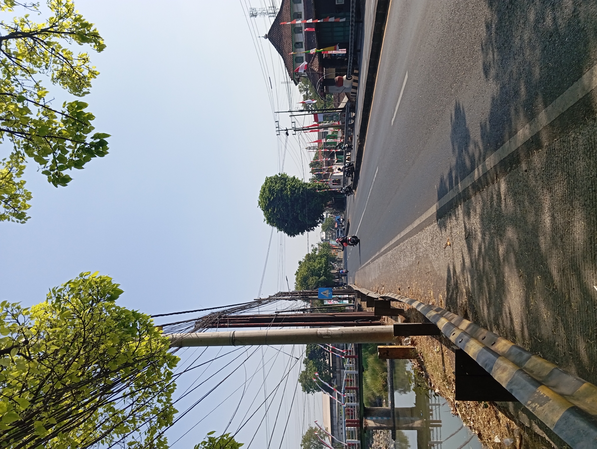 This is a daytime, eye-level shot of a street scene in Kepulungan, Indonesia. The image is dominated by a two-lane asphalt road curving to the right. On the left side of the road, there is a concrete sidewalk bordered by a yellow and black striped guardrail. Beyond the guardrail, a body of water with some debris on its banks is visible, with a bridge spanning it.
Several utility poles with a dense network of wires are positioned along the sidewalk. A few trees with bright green leaves overhang the scene from the top left and right corners.
On the road, two motorcycles are traveling in the same direction. One is closer to the foreground and ridden by a person wearing a helmet and a jacket. Another motorcycle is further down the road.
The right side of the road features buildings with traditional Indonesian architecture, adorned with numerous red and white flags. One building appears to be a mosque with a distinctive domed roof. In the background, more trees and utility towers can be seen against a clear blue sky. The overall atmosphere suggests a sunny day in a semi-urban environment.