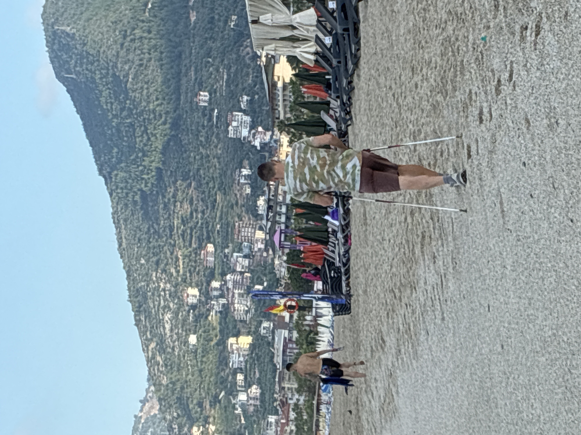 The image depicts a beach scene in Alanya, Turkey. In the foreground, a man with a camouflage-patterned t-shirt and brown shorts is walking on the sand, using Nordic walking poles. Behind him, another man, shirtless and wearing dark swim trunks with a colorful pattern, is walking away from the camera, carrying what appears to be a blue towel or shirt.
The beach is lined with numerous lounge chairs and umbrellas, indicating it is a developed tourist area. In the background, the hills of Alanya are visible, dotted with white buildings and interspersed with green vegetation. The sky is clear and blue. The overall impression is a summer day at a coastal resort.
