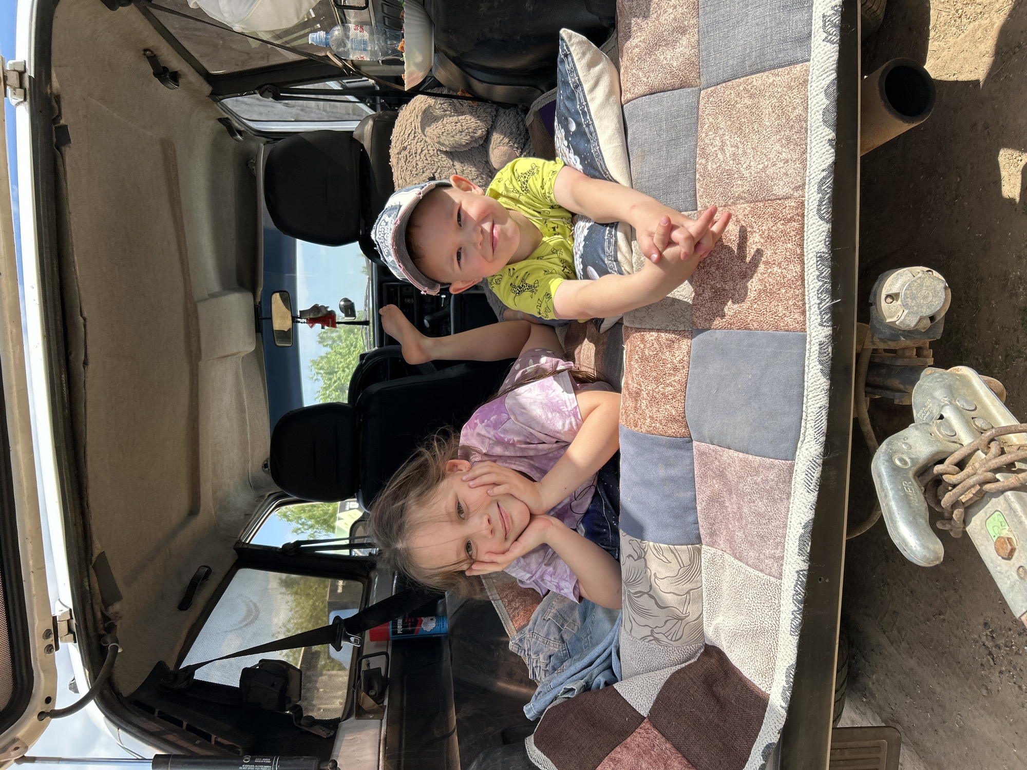 Two young children, a boy and a girl, are lying in the back of a vehicle, possibly a van or SUV. The interior of the vehicle is visible, including the back seats, headrests, and rear window. The vehicle is located in Фоки, Russia, as indicated in the location data.
The boy is wearing a yellow shirt with a giraffe pattern and a baseball cap. He is lying on his back with his hands clasped together. The girl is wearing a purple tie-dye shirt and is lying on her side with her head resting on her hands.
They are lying on a patchwork quilt or blanket that is covering the cargo area of the vehicle. A pillow and a stuffed animal are also present. There are various items stowed in the front of the vehicle. Outside the vehicle, a tow hitch is visible.