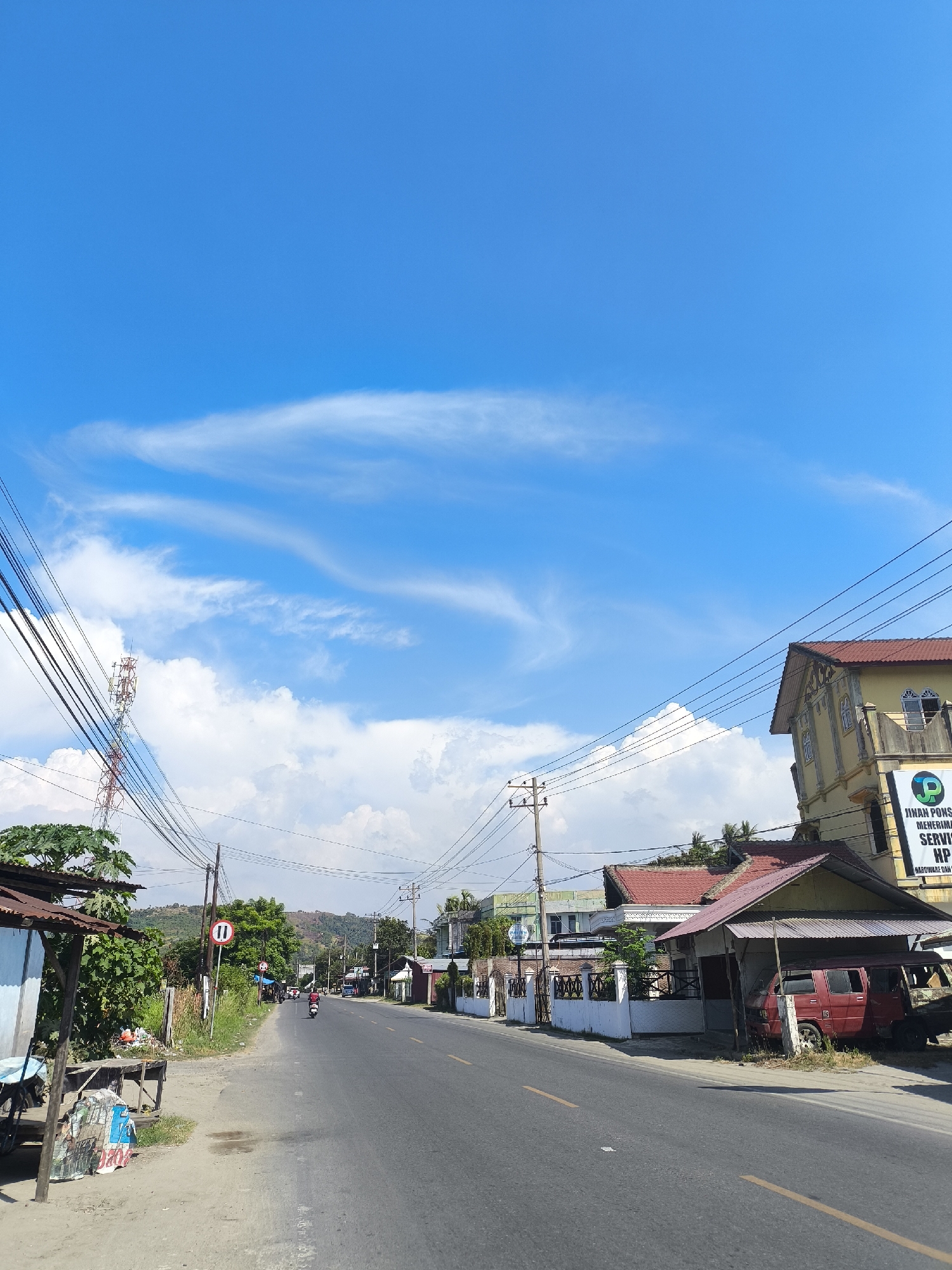 The image depicts a street scene in Juliseutuy, Indonesia. A paved road with yellow lane markings stretches into the distance, bordered by buildings and vegetation. The sky is bright blue with scattered white clouds. On the left side of the road, there is a small, rustic stall with a corrugated metal roof, a wooden structure, and a parked motorcycle. Further down the road, traffic signs are visible, along with a telecommunications tower. On the right side of the road, a multi-story building with a yellow facade and a red-tiled roof stands prominently. Another building with a red-tiled roof and an open-fronted structure housing a red van is situated next to it. A banner on the yellow building advertises "JIHAN PONSEL" and "MENERIMA SERVIS HP HARDWARE DAN SOFTWARE." A few motorcycles are seen on the road, suggesting a moderate level of activity. The background shows rolling hills and trees under a clear sky.