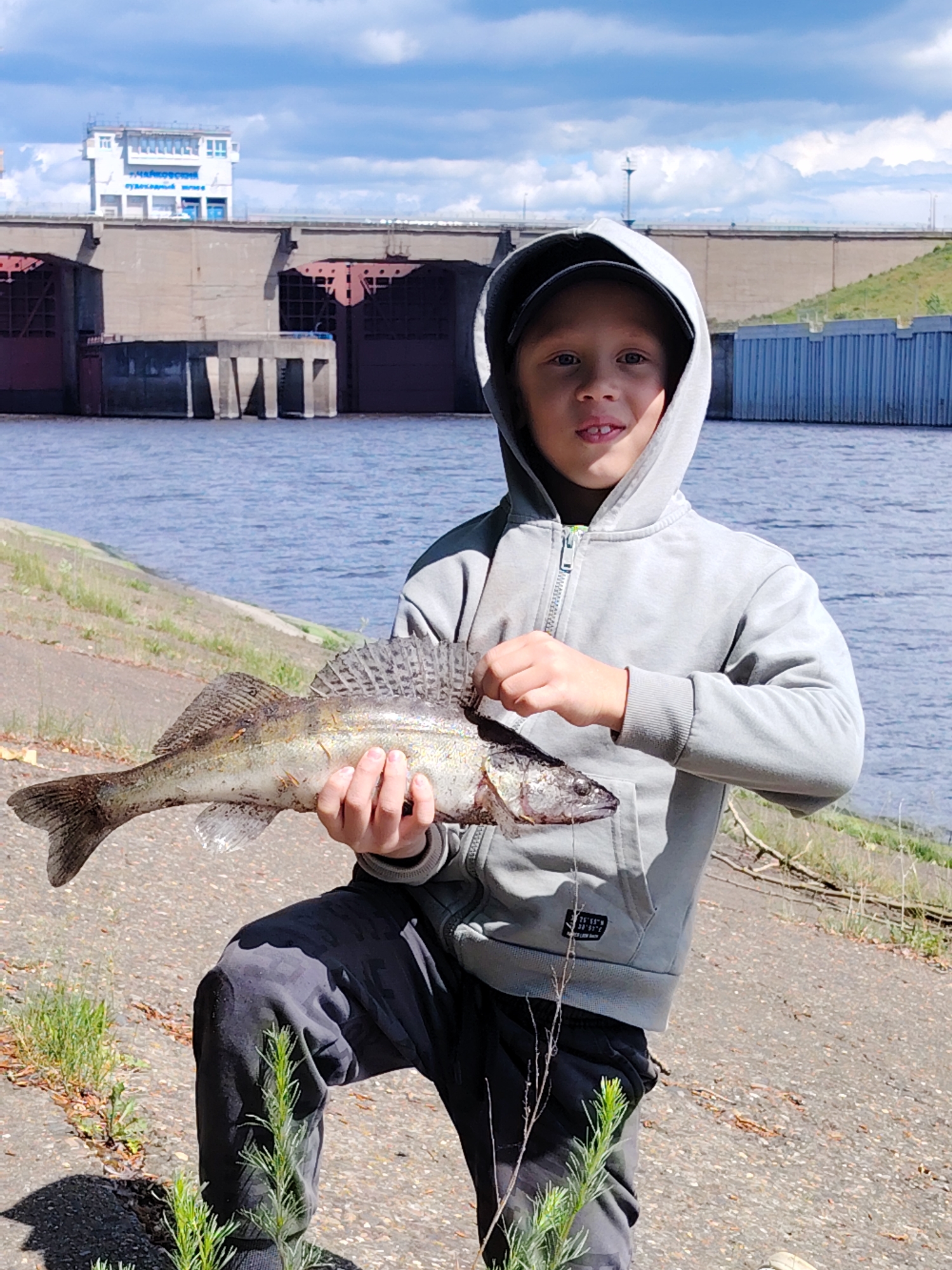 A young boy holds a large fish in front of a dam. The boy is wearing a gray hooded sweatshirt and gray camouflage pants. He is kneeling on a concrete surface with grass and weeds growing in the cracks. In the background is a large dam with the name "Votkinsk" in Russian written on the control tower. The water in the foreground is part of the Kama River in Chaikovsky, Russia.