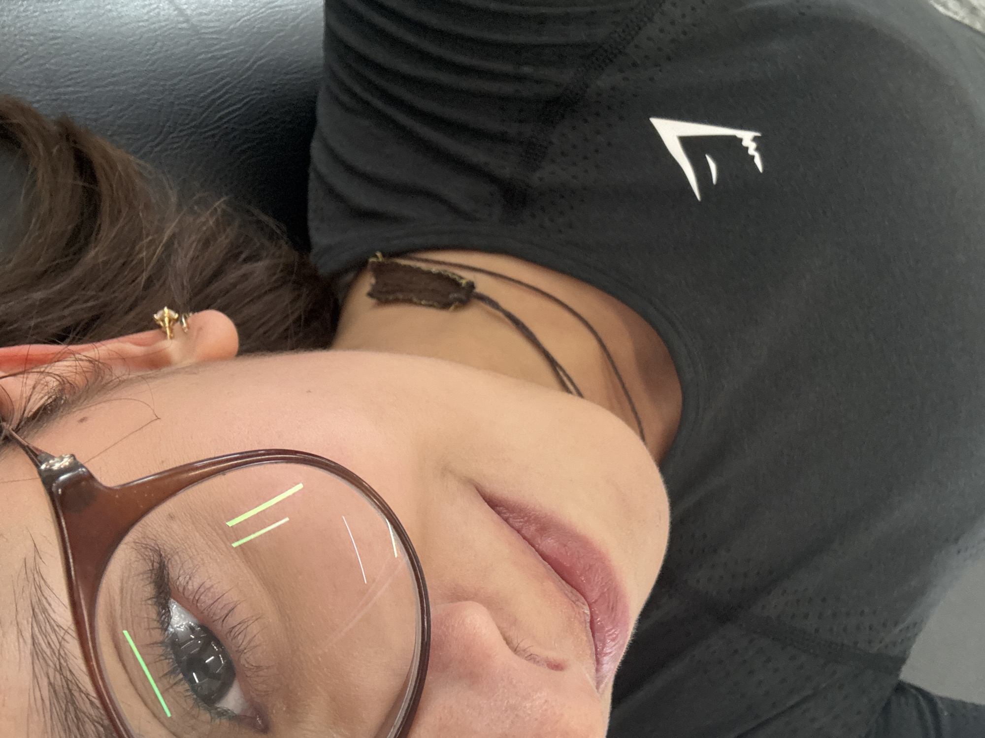 A close-up, high-angle shot captures a person, likely female, from the chest up, positioned on their side in what appears to be an indoor setting in San Andrés Cholula, Mexico. The subject is wearing brown-framed glasses, which reflect light as green and white linear streaks across the lenses. Their left eye, with long eyelashes, is visible through the lens. A small, gold-colored earring, possibly with a tiny clear stone, is present in the upper part of their left ear, adorned with dark brown, wavy hair. The person's lips are slightly parted, showing a muted pink lipstick or natural lip color. Around their neck, a dark, flat strap, possibly made of leather or fabric, is visible, leading to a dark brown, rectangular element behind the neck. They are dressed in a black athletic top made of a perforated, breathable material. A distinctive white, angular logo, identified as the Gymshark logo, is prominently displayed on the front of the top. The background is a plain, dark color, suggesting upholstery or a soft surface. The overall impression is one of casual relaxation, with the person likely resting or posing for a close-up photo.