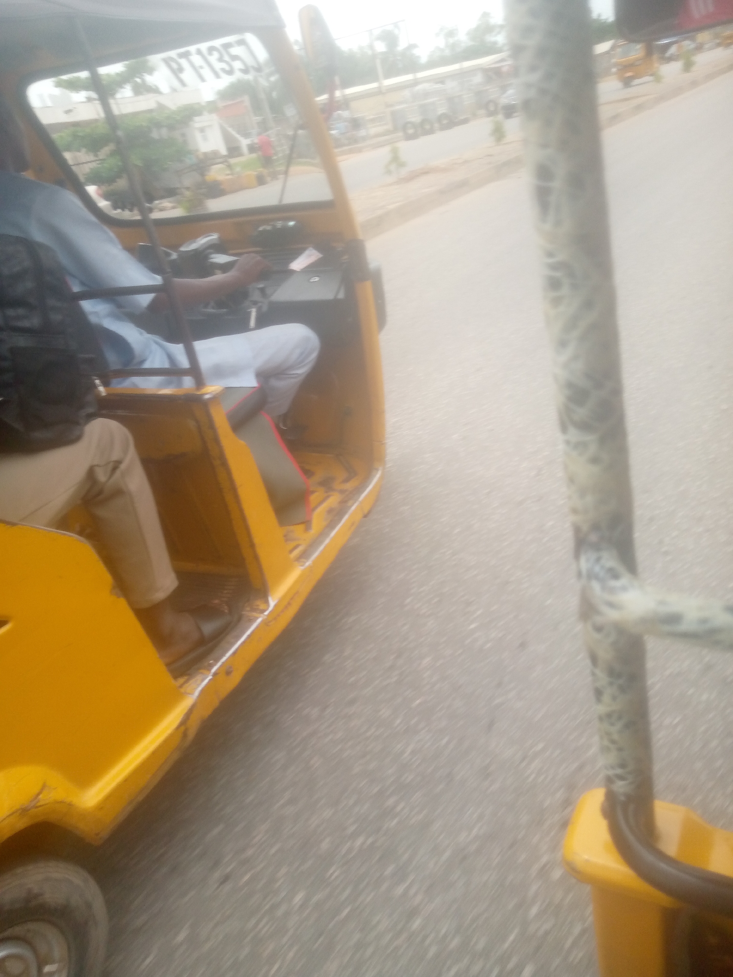 The image shows the interior of a yellow auto-rickshaw. A person wearing a light blue shirt and white trousers is driving the auto-rickshaw, with their hands on the steering wheel. Another person, wearing a dark jacket and beige trousers, is seated to the left. The auto-rickshaw is on a paved road in what appears to be an urban environment. In the background, buildings and other vehicles are visible. The text "PT135J" is visible on the windshield of the auto-rickshaw. The image seems to be taken from the perspective of a passenger in the same auto-rickshaw, looking forward and slightly to the right. The environment suggests a location in Jalingo, Nigeria, given the context provided.