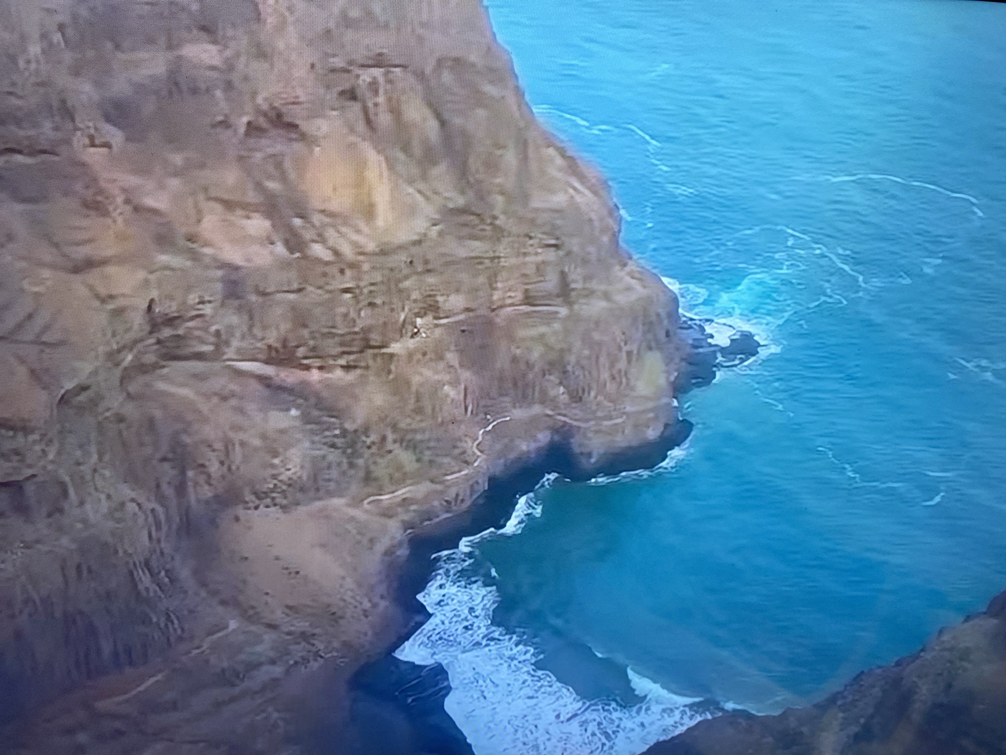 The image shows a high-angle view of a rugged, rocky cliff face meeting a bright turquoise ocean. The cliff is textured with layers of brown and beige rock, exhibiting signs of erosion and weathering. The ocean displays white-capped waves crashing against the base of the cliff, creating a foamy white border where the water meets the land. The water itself is a vibrant blue, with subtle variations in color suggesting depth. There are no visible people, structures, or any indications of the city of Minsk, Belarus, in this scene. The environment appears to be a natural coastal landscape.