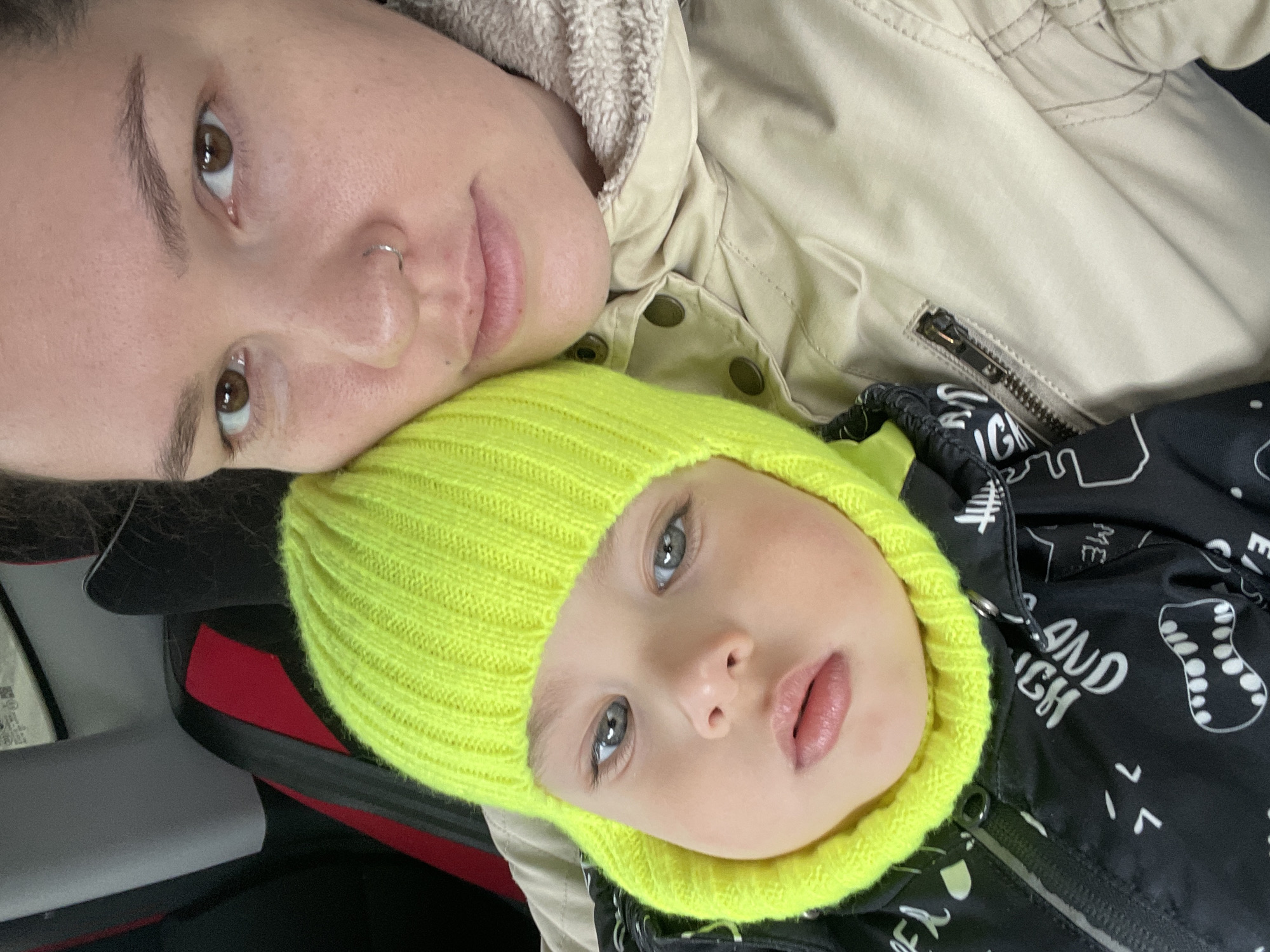 The image shows a woman and a child lying down. The woman has brown hair and eyes and a nose ring. She is wearing a beige jacket. The child is wearing a bright yellow knit hat and a black jacket with white designs. The background suggests they are in a car, specifically a car seat with red accents. There is no specific information in the image indicating it is in Izhevsk, Russia.