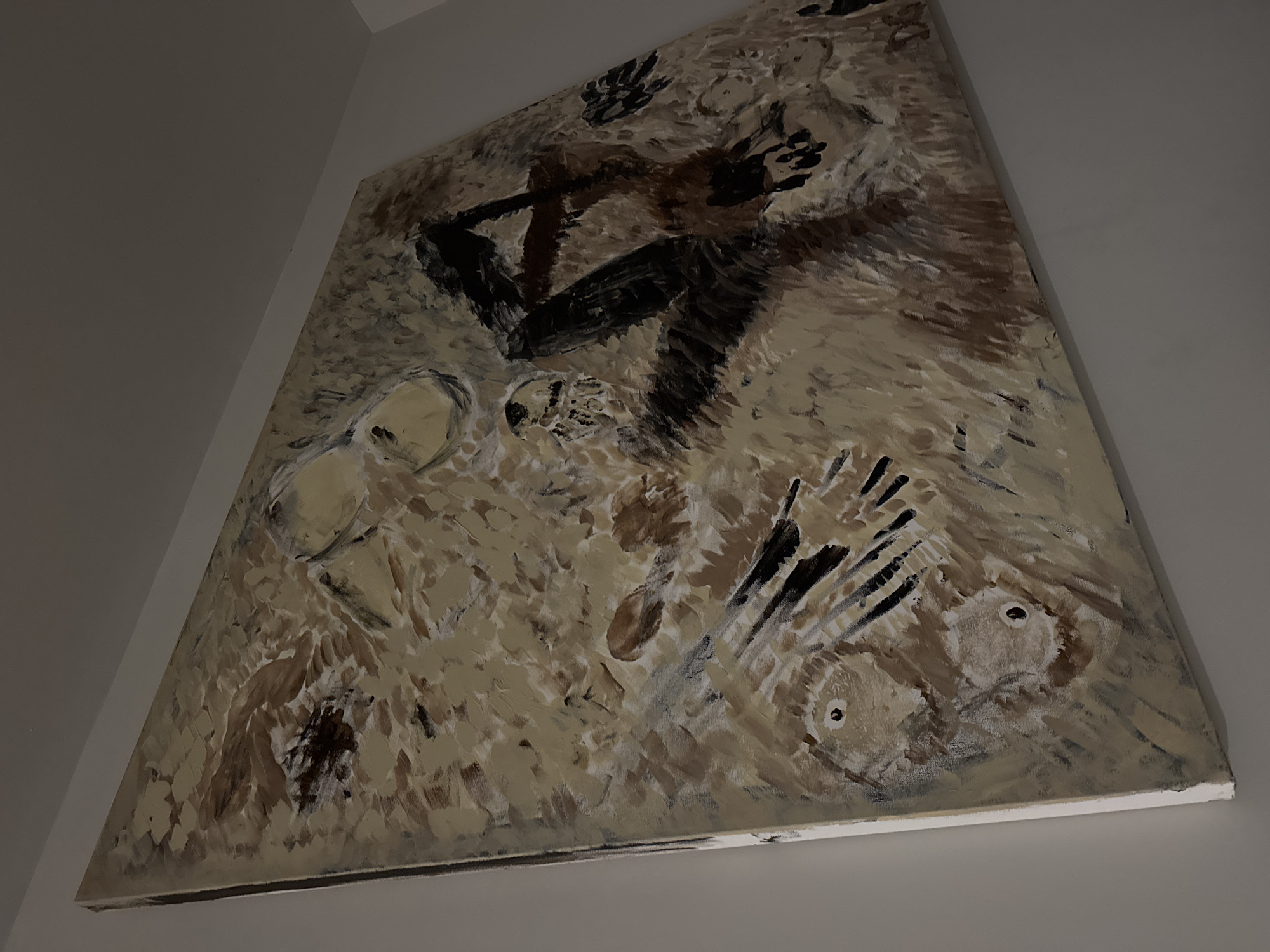A large, abstract painting hangs on a white wall. The artwork is rendered in shades of brown, beige, and white, with bold black accents. The painting is positioned in the city of Unknown, Unknown. Recognizable shapes include human figures with outstretched arms. Additional shapes appear as skull-like forms. The overall composition is textured and gestural.