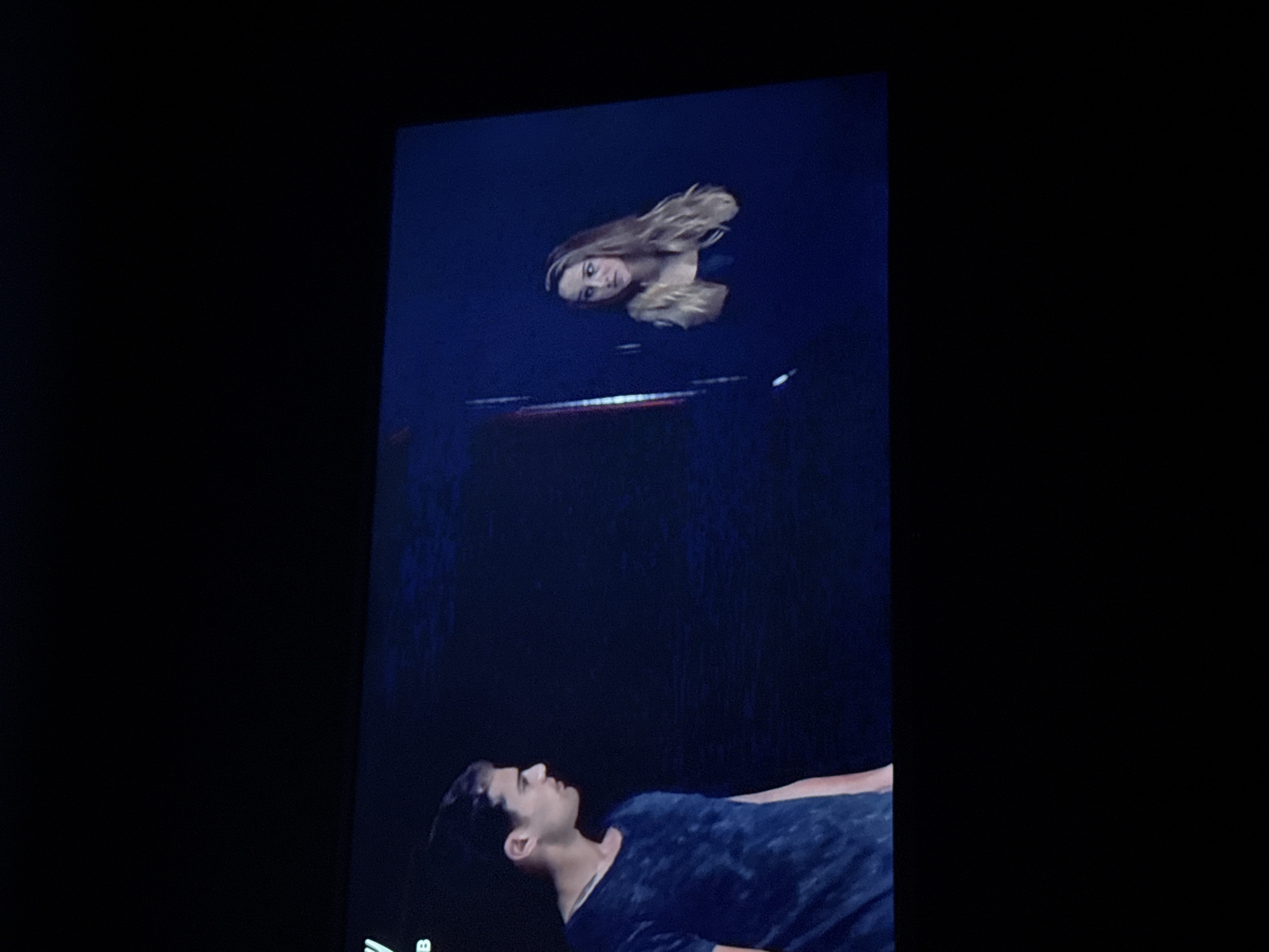 The image depicts a scene from a screen, potentially a television or monitor. At the top of the screen is a woman with long blonde hair, appearing to be floating in a dark space, possibly water. Her head is tilted slightly to the left, and she is looking down. Below her, there is a blurred, reddish-white horizontal object. At the bottom of the screen is a man lying down, looking up towards the woman. He has dark hair and is wearing a dark t-shirt. The surrounding environment is black, indicating a dark room or background. The image does not contain any information to determine if this scene is related to Mytishchi, Russia.