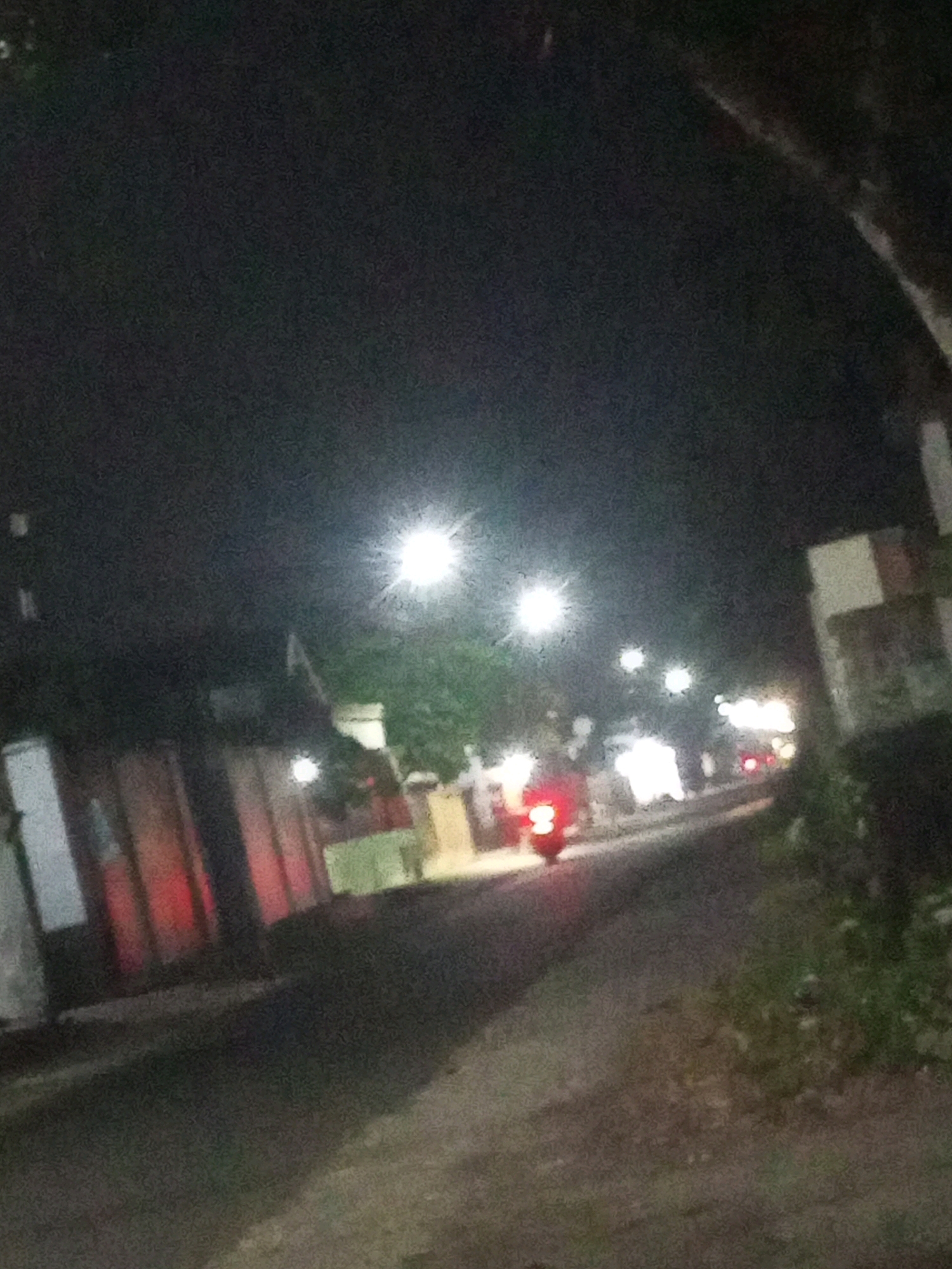 The image depicts a street scene at night in Ngasem, Indonesia. A motorcycle, illuminated by its red taillight and brake light, is moving away from the viewer on the left side of the asphalt road. The road is bordered by buildings on the left and vegetation on the right. Streetlights are visible, casting a bright glow in the dark environment. The overall impression is that of a typical residential or commercial street in a dimly lit area at night.