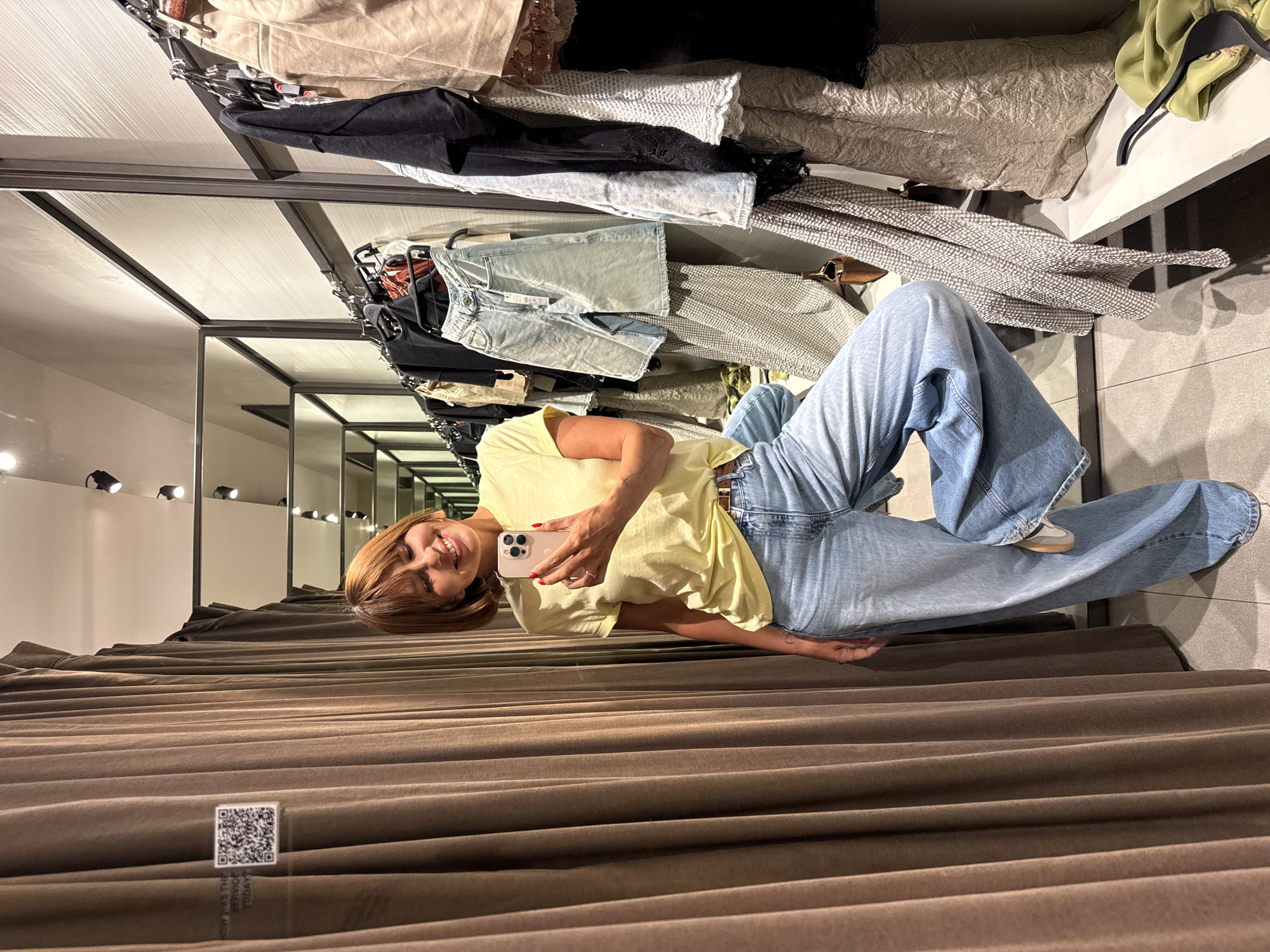 The image, taken in Kuta, Indonesia, captures a woman taking a selfie inside a clothing store fitting room. The perspective is rotated 90 degrees counter-clockwise, making the woman appear to be lying down on the curtain.
The central subject is a woman with light brown, shoulder-length hair, wearing a light yellow short-sleeved top, light blue wide-leg denim jeans, and light-colored sneakers with a brown accent. She holds a light-colored smartphone in her right hand, positioned to take a selfie, and is smiling broadly at the camera. Her fingernails are painted red.
The setting is a well-lit changing room. In the background (which would be to her right and above if the image were correctly oriented), clothing racks are visible, holding various garments including denim shorts, light fabrics, and darker tops. Multiple mirrors reflect parts of the room and overhead spotlights. The foreground and the side closest to her face are dominated by a dark brown, vertically ridged fitting room curtain, pulled shut to create a private space.
A small, clear sign with a black QR code is attached to the curtain in the lower left corner. Partially visible text beneath the QR code reads "HOW WAS THE SERVICE TODAY?". The time of day is not discernible due to the indoor artificial lighting, and the woman's smile suggests a pleasant or playful mood.